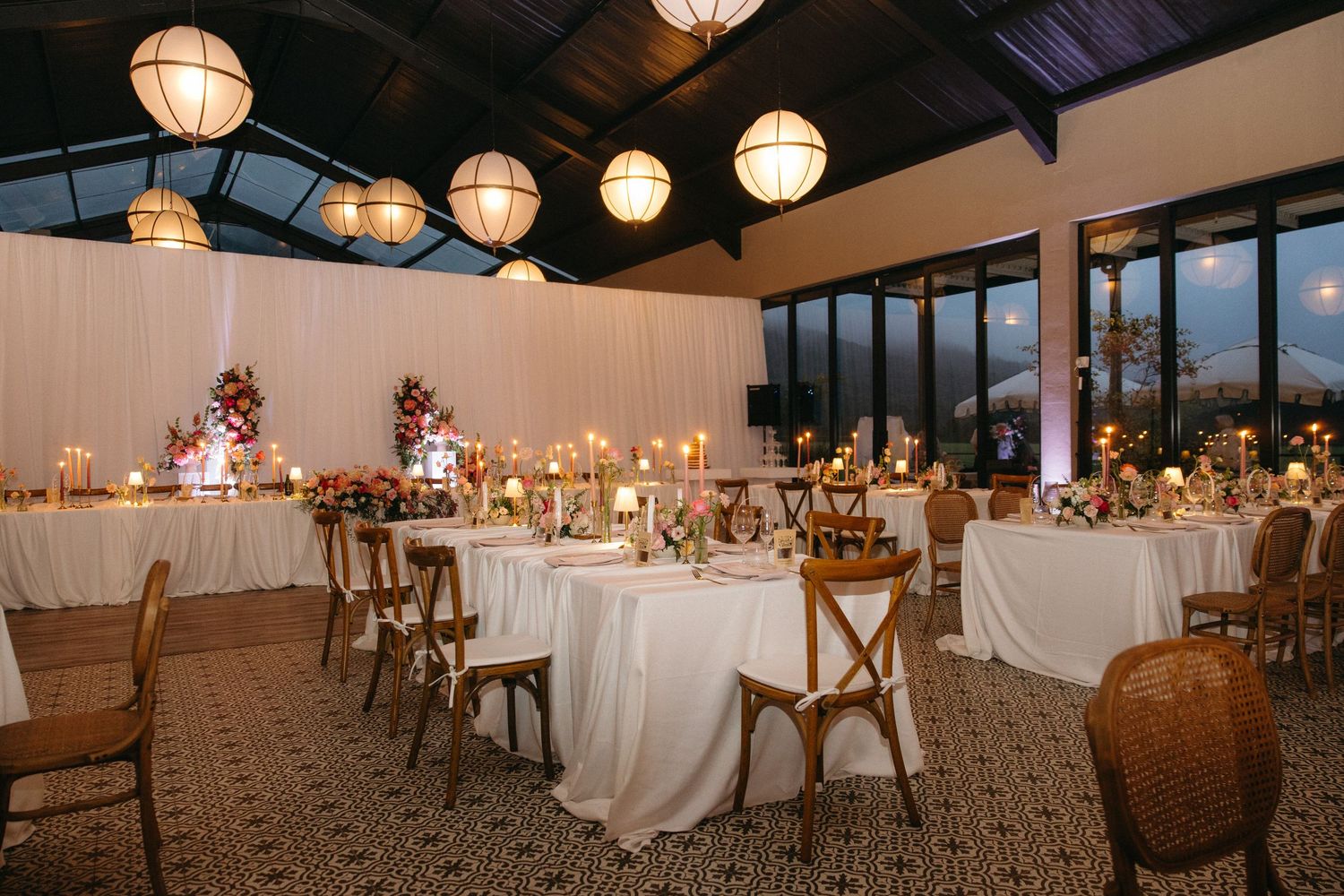 Elegant wedding reception hall with white draped walls, round tables, candlelight, and woven rattan chairs under hanging lanterns.
