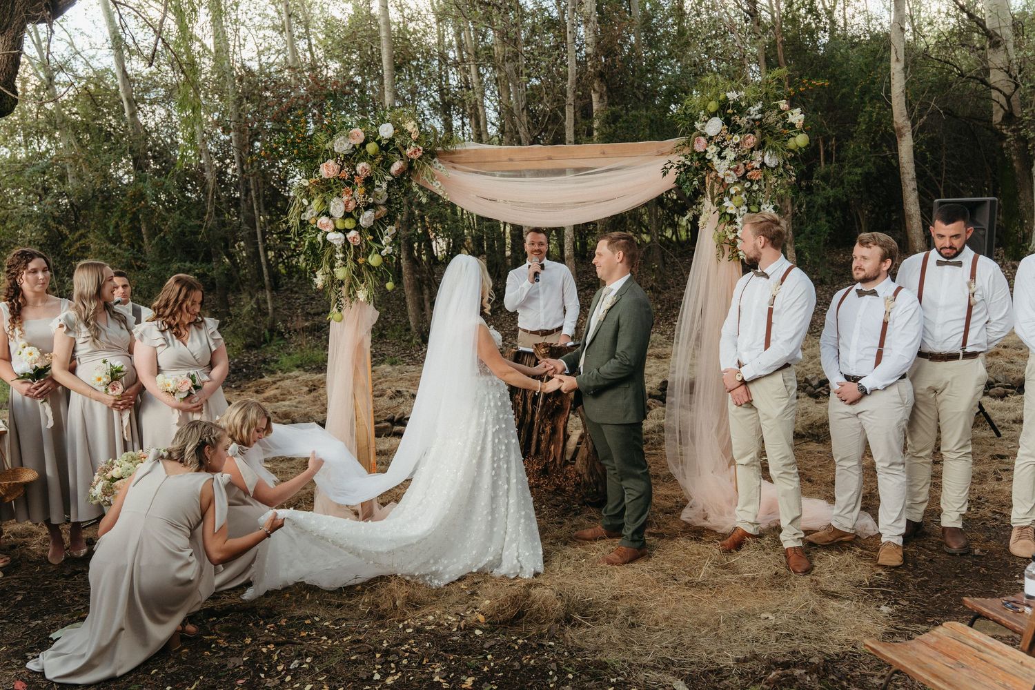 Outdoor woodland wedding ceremony with bride and groom under floral arch, bridesmaids in gray, groomsmen in suspenders.