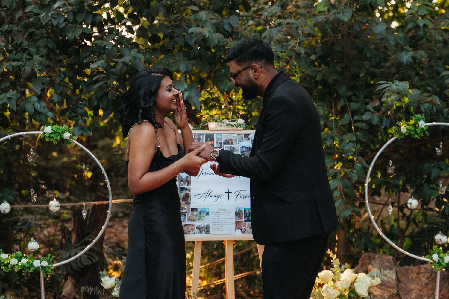 Couple sharing a joyful moment at an outdoor engagement setup with floral decor and a personalized photo display board.