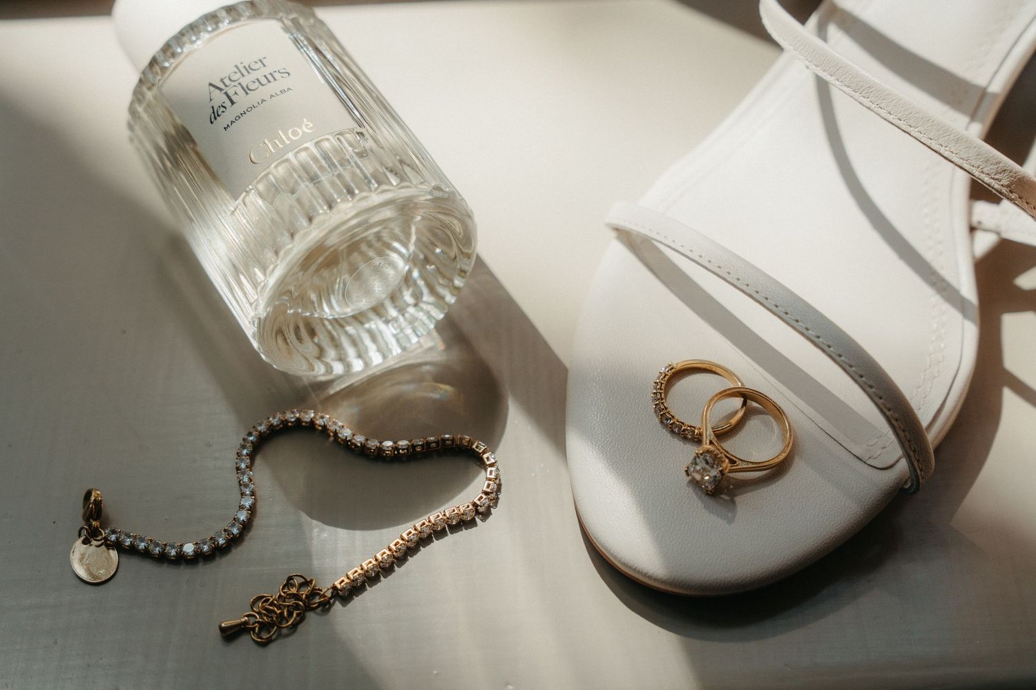 Elegant bridal flat lay with gold rings on white heels, pearl bracelet, key charm, and glass perfume bottle in soft sunlight.
