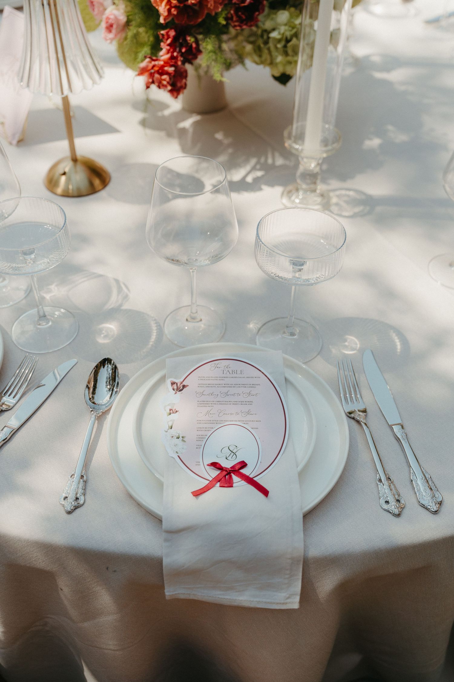 Elegant outdoor wedding table setting with white plates, menu card, red ribbon, crystal glasses, and floral centerpiece on white linen.