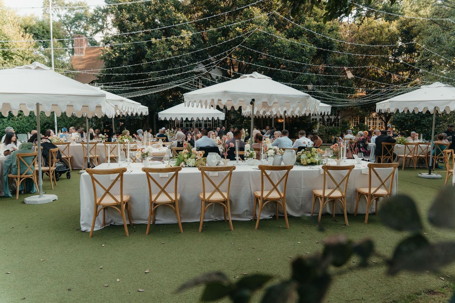 Elegant outdoor wedding reception with white marquee tents, string lights, and wooden cross-back chairs on a green lawn.