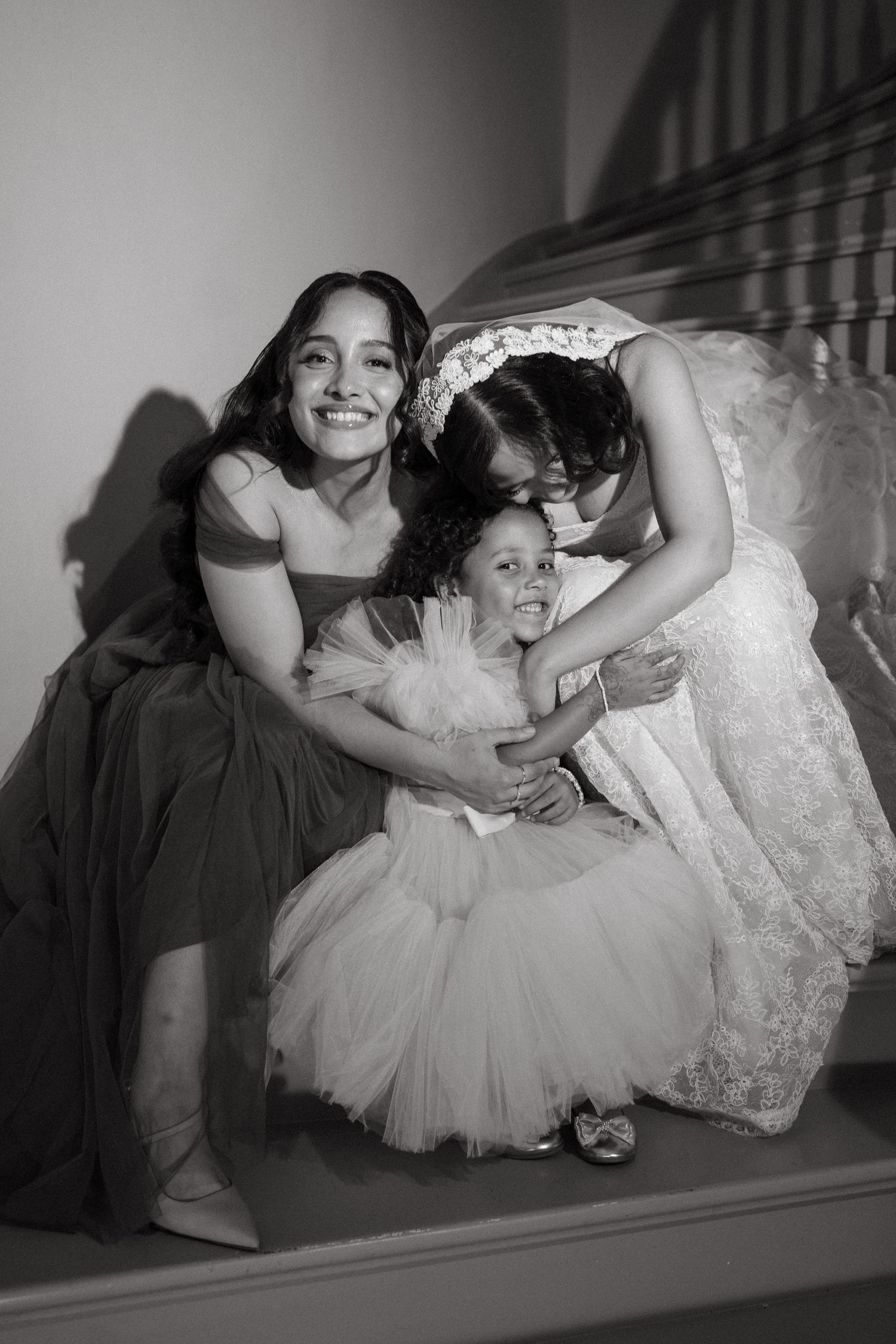 Black and white photo of a bride in lace gown hugging a flower girl and bridesmaid on wedding stairs.