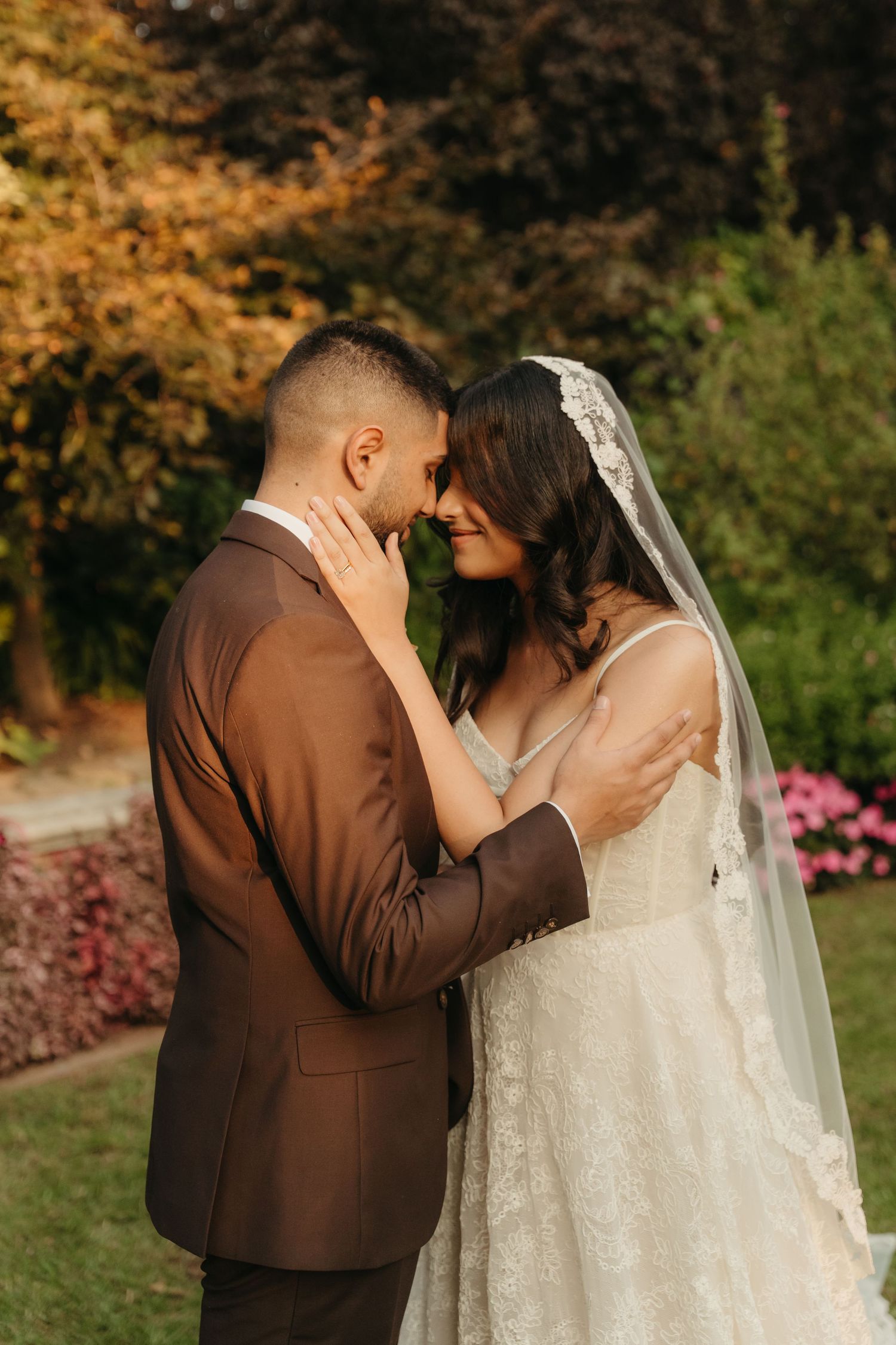 A bride and groom share a romantic forehead touch in a lush garden, she in a lace gown with veil, he in a brown suit.
