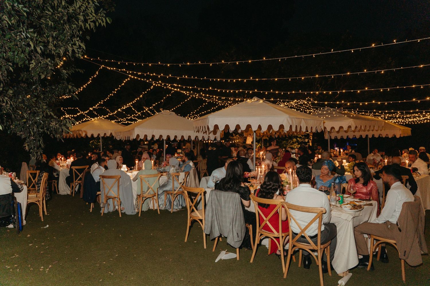 Outdoor evening wedding reception with string lights, white tent, and guests dining at candlelit tables under the night sky.