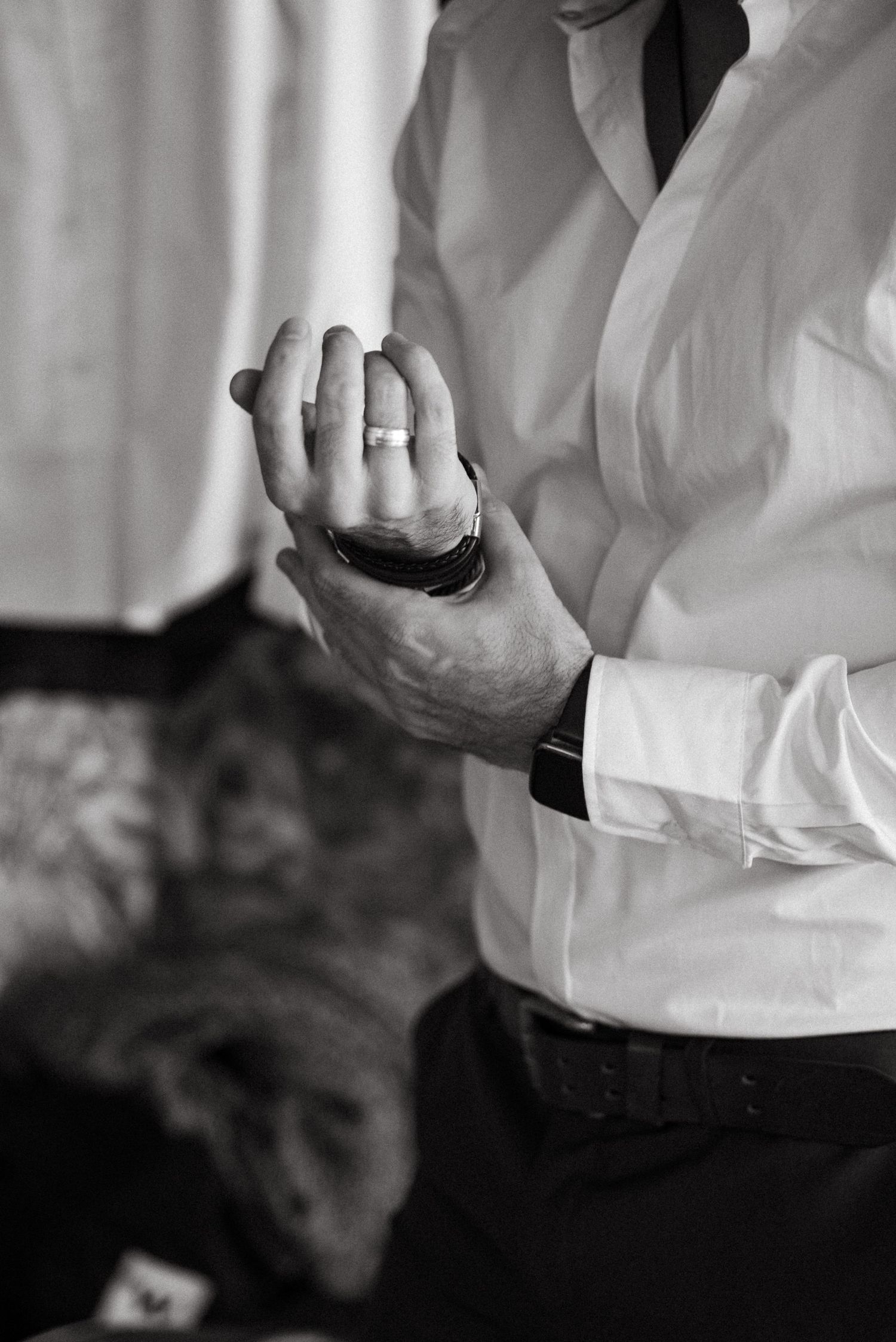 Black and white photo of a groom in white dress shirt adjusting cufflinks, wearing a wedding band.