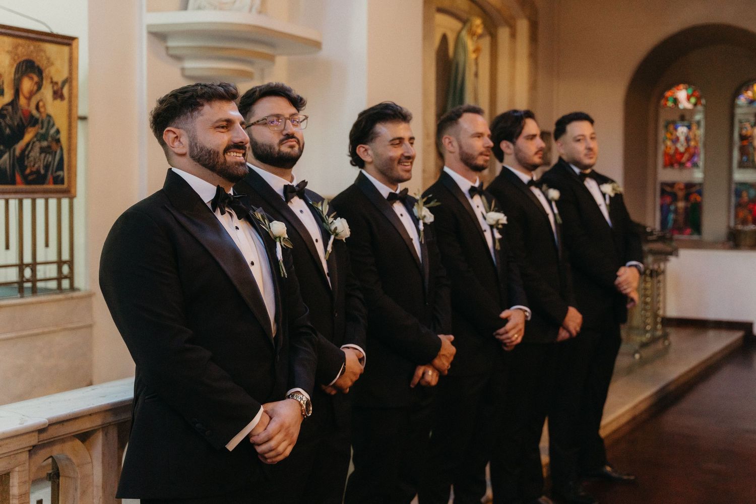 Groom and groomsmen in black tuxedos with bow ties and boutonnieres standing at Rosebank Catholic Church altar during wedding ceremony.