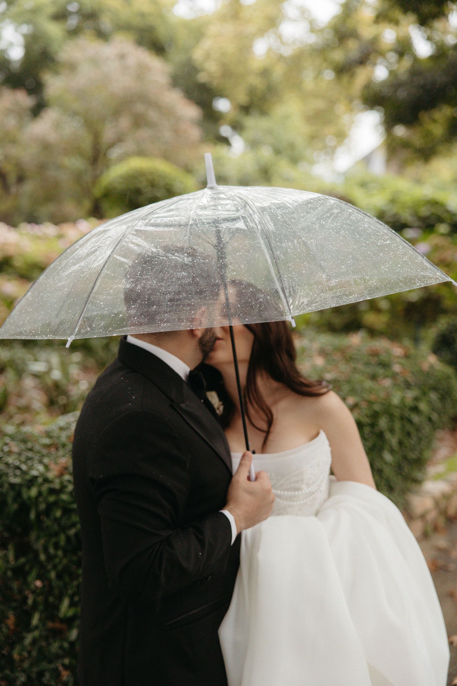 Bride and groom sharing a kiss under a clear umbrella in a lush garden on a rainy wedding day.