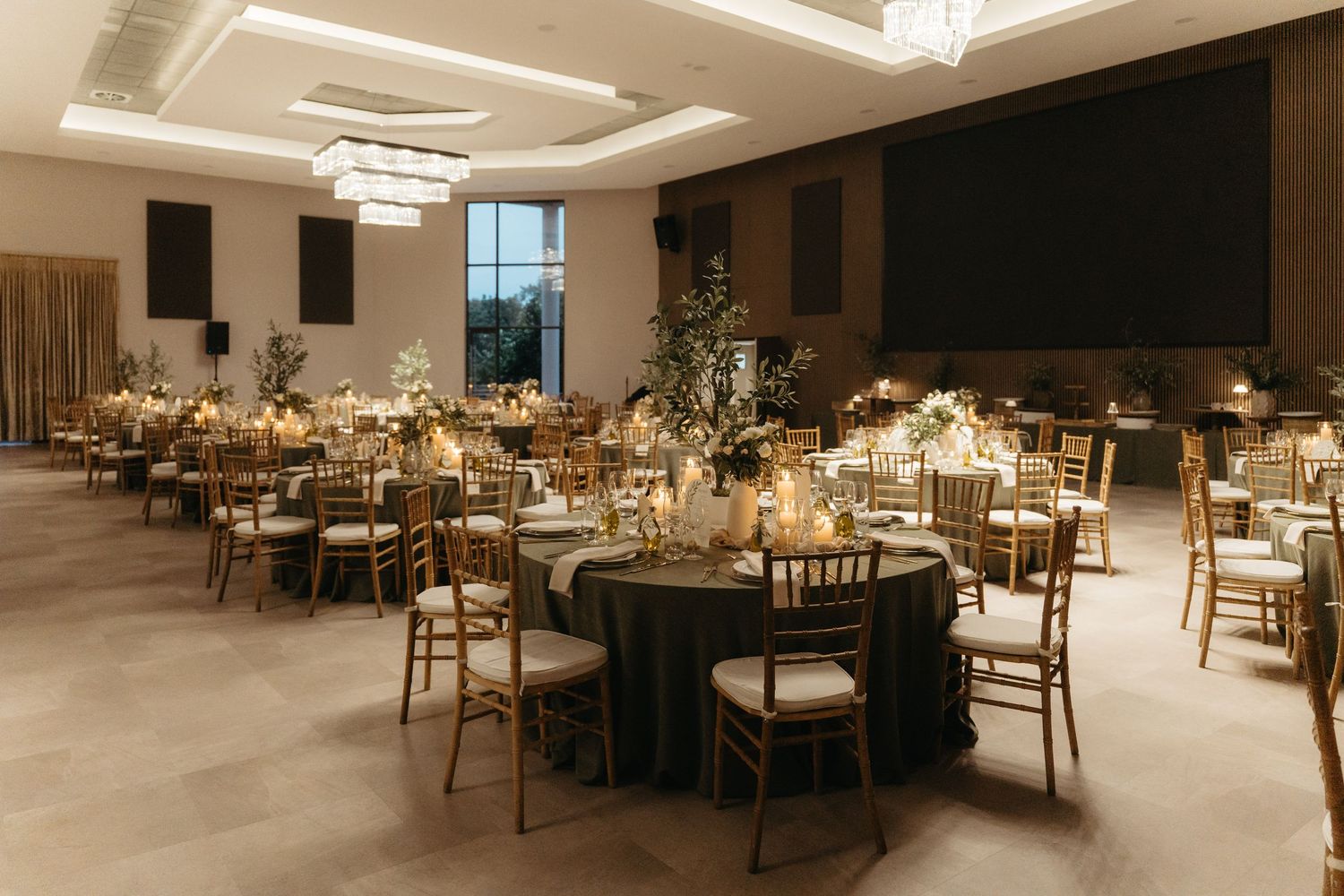 Elegant wedding reception hall with round tables, gold Chiavari chairs, dark linens, floral centerpieces, and warm candlelight.