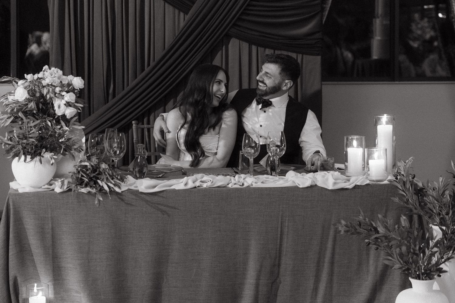 Bride and groom share a laugh at candlelit sweetheart table during wedding reception.