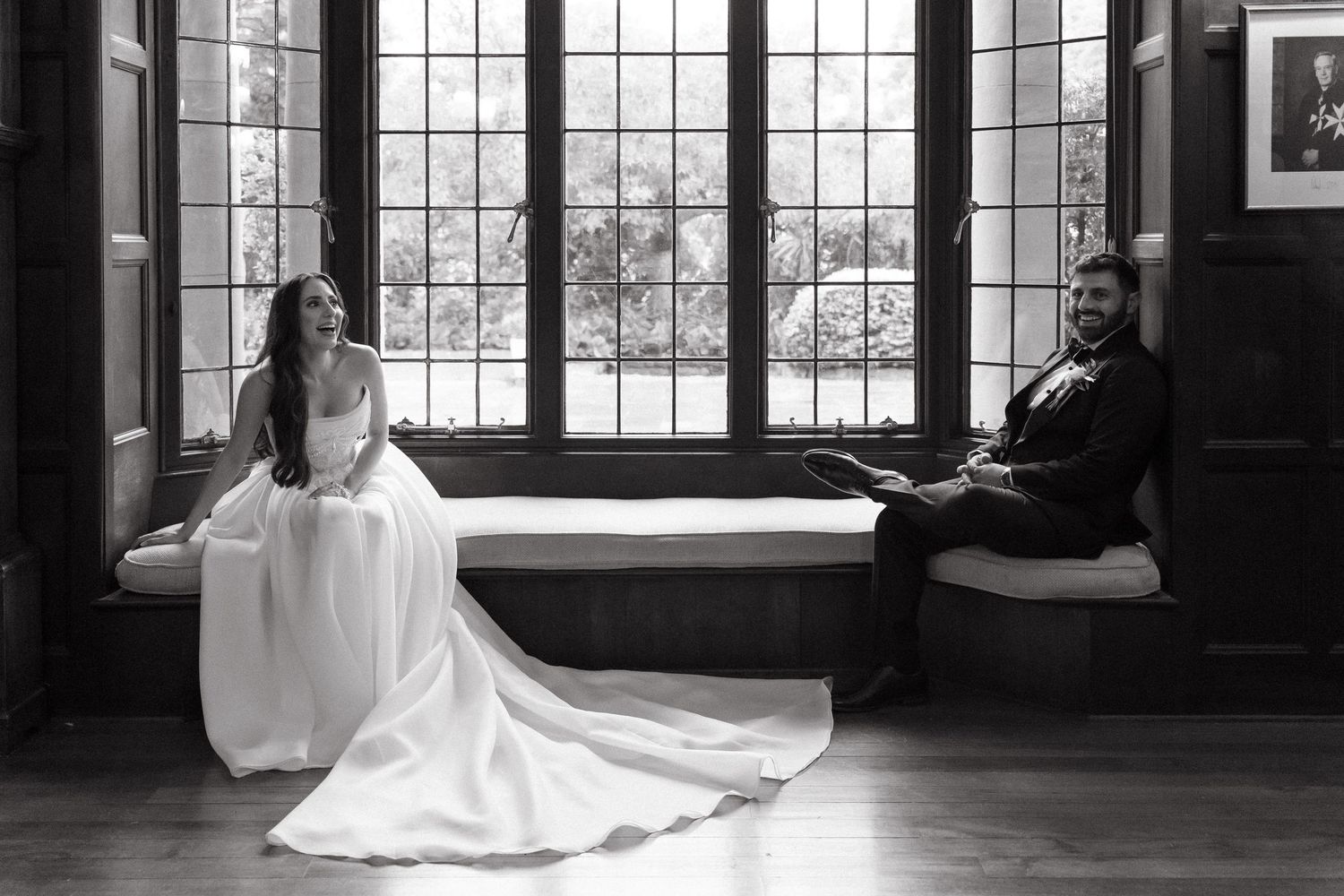 Black and white wedding photo at Glenshiel of bride and groom sitting apart on window bench, smiling in elegant attire.
