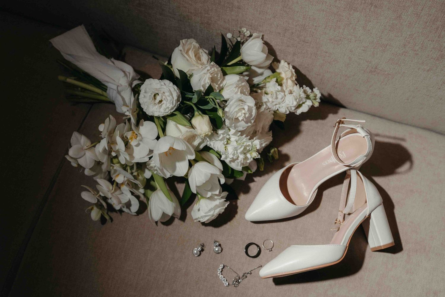 Elegant bridal flat lay featuring white floral bouquet, cream block heel shoes, and delicate rings on a neutral background.