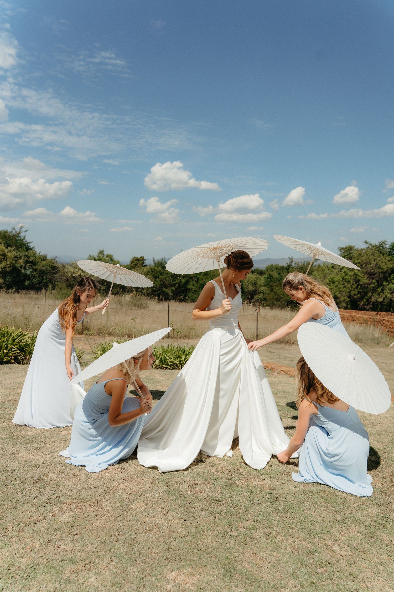 Bride in white gown with bridesmaids in blue dresses holding parasol umbrellas outdoors at Steynshoop Valley Lodge