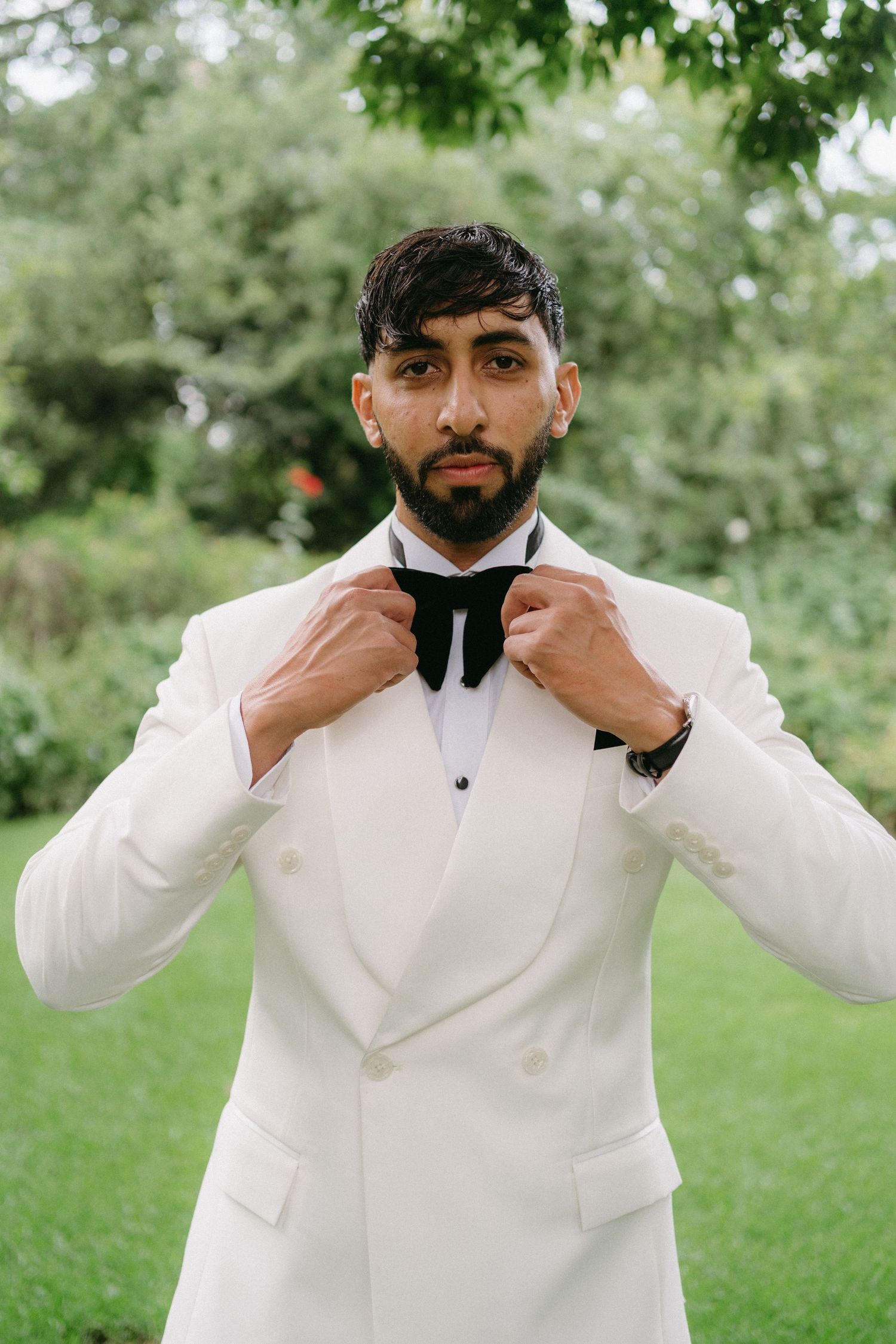 A formal portrait of someone adjusting their bowtie while wearing a crisp white tuxedo jacket against a green outdoor backdrop.