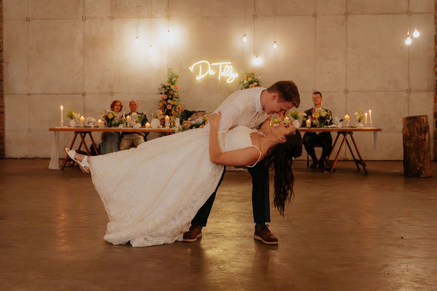A couple shares a romantic dance in a rustic venue decorated with twinkling string lights overhead.