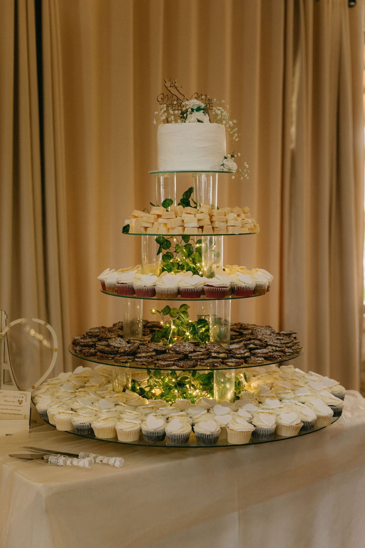 Elegant tiered dessert display with cupcakes and treats illuminated by string lights.