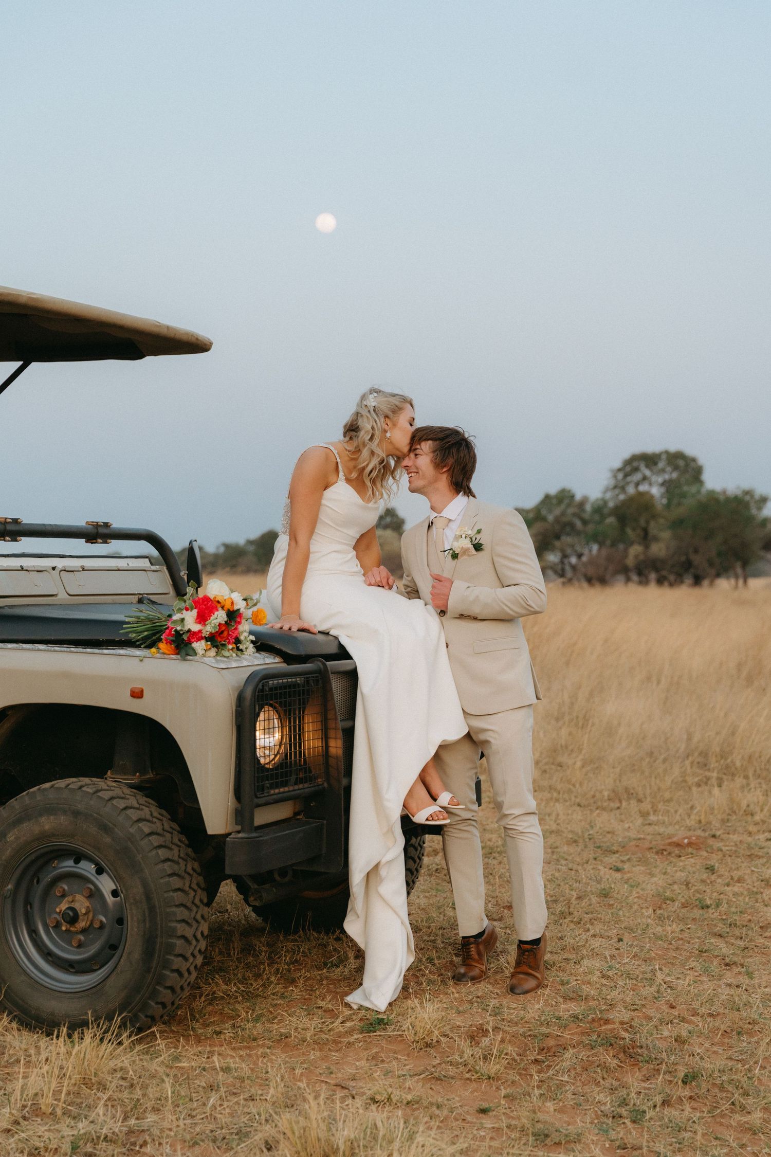 Romantic wedding photo of a couple by a vintage Land Rover at dusk.
