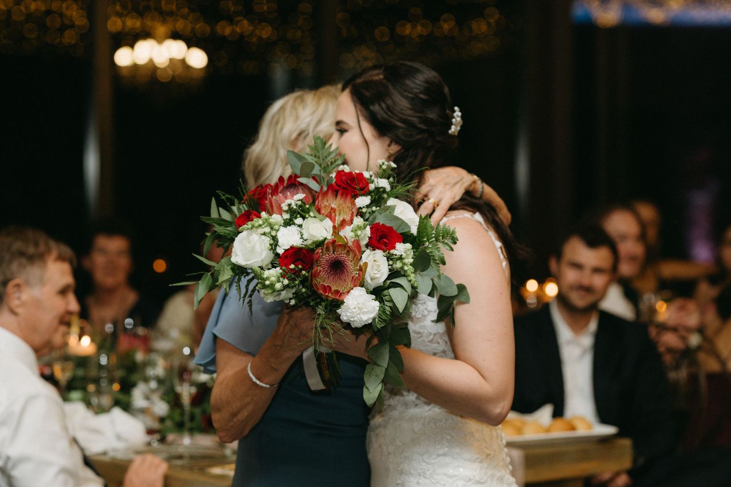 Two people embrace at a wedding reception while holding a bouquet of red and white flowers.