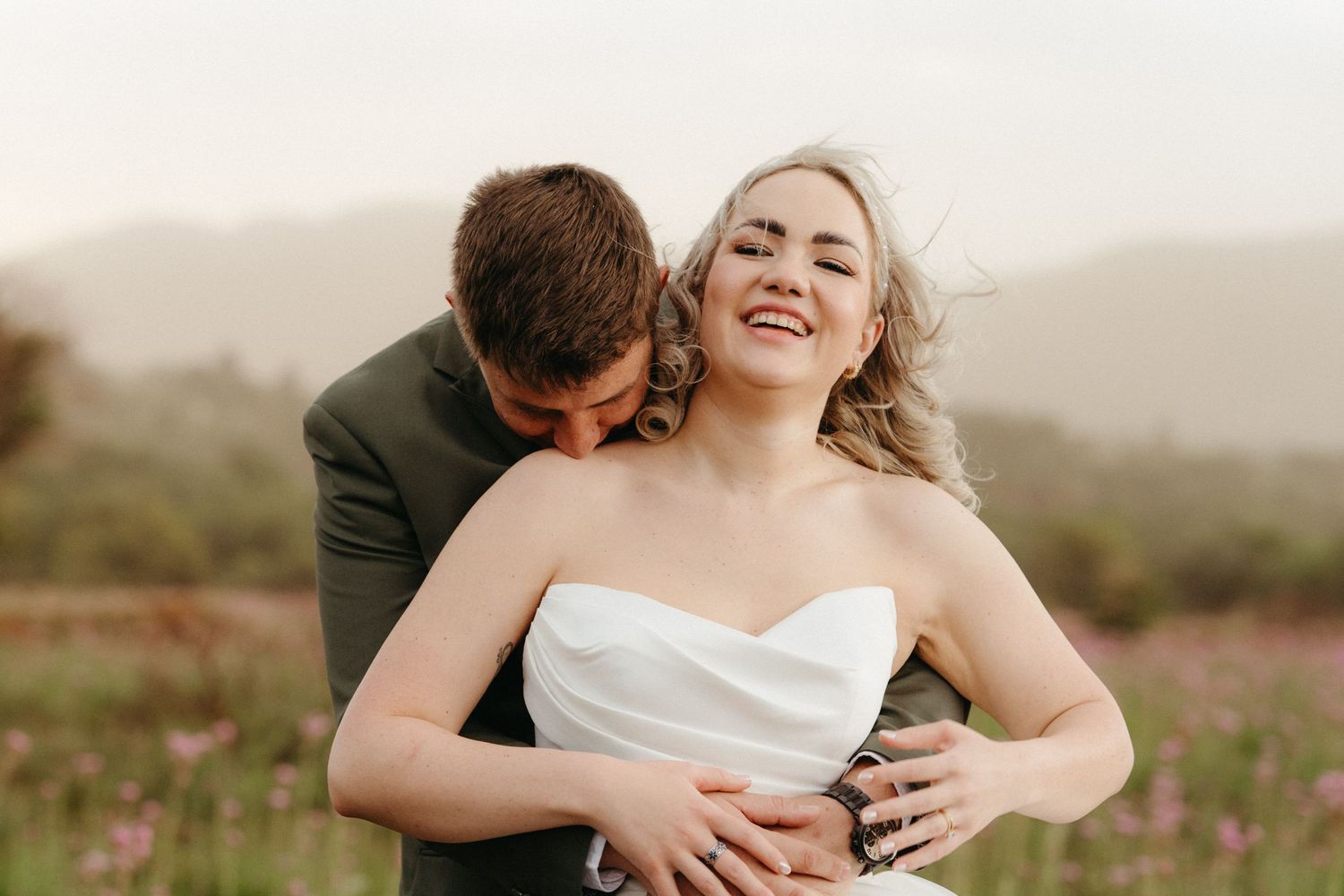 A romantic wedding couple embraces in a sunlit meadow with rolling hills in the background during golden hour.