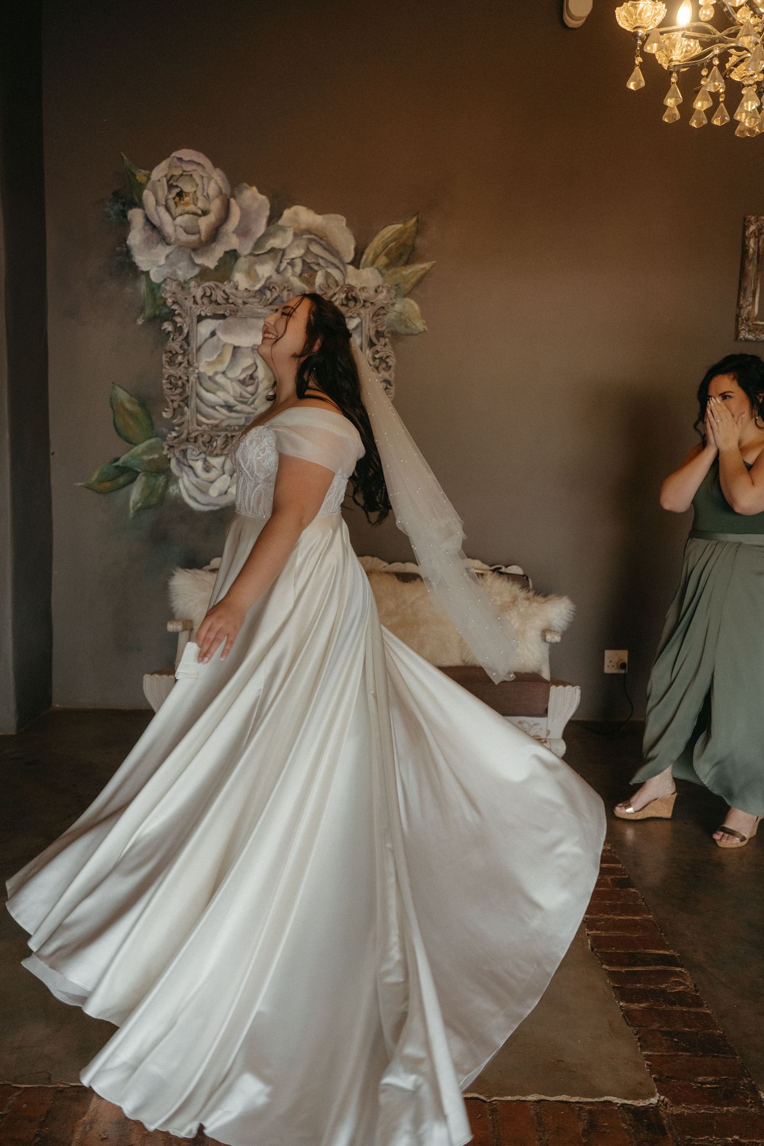 Bride in flowing white dress twirling gracefully in front of floral wall decor.