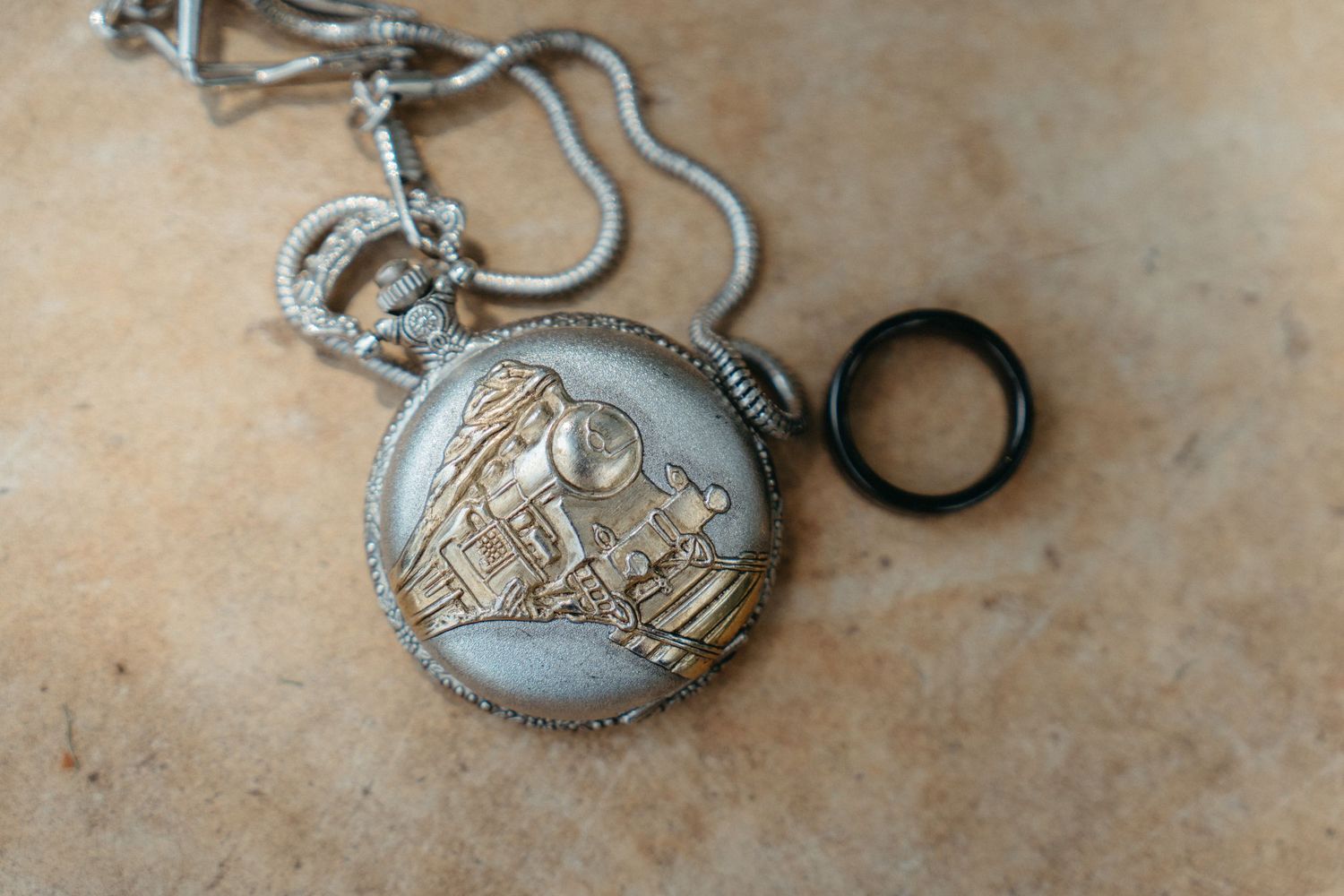 Ornate silver pocket watch with decorative engraving and chain displayed on textured surface.