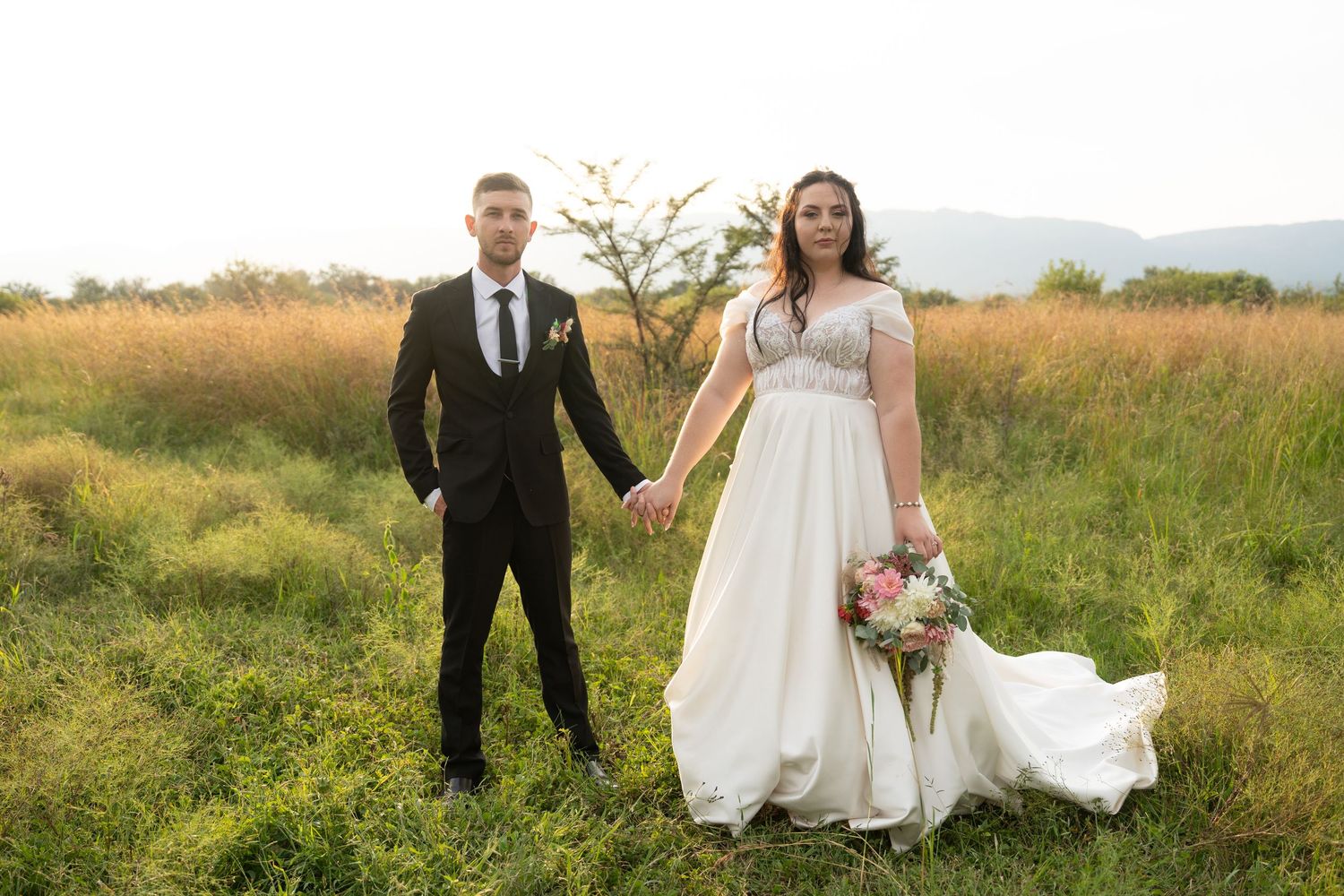 Newlyweds pose together in a serene field with mountains in the background.
