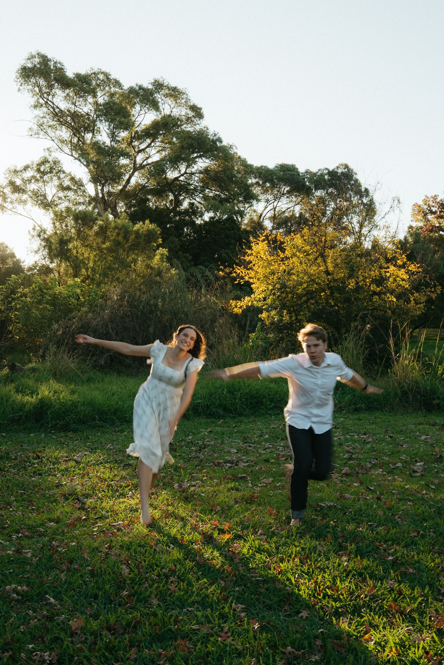 Playful couple dances joyfully in a grassy field with golden autumn trees in the background at sunset.