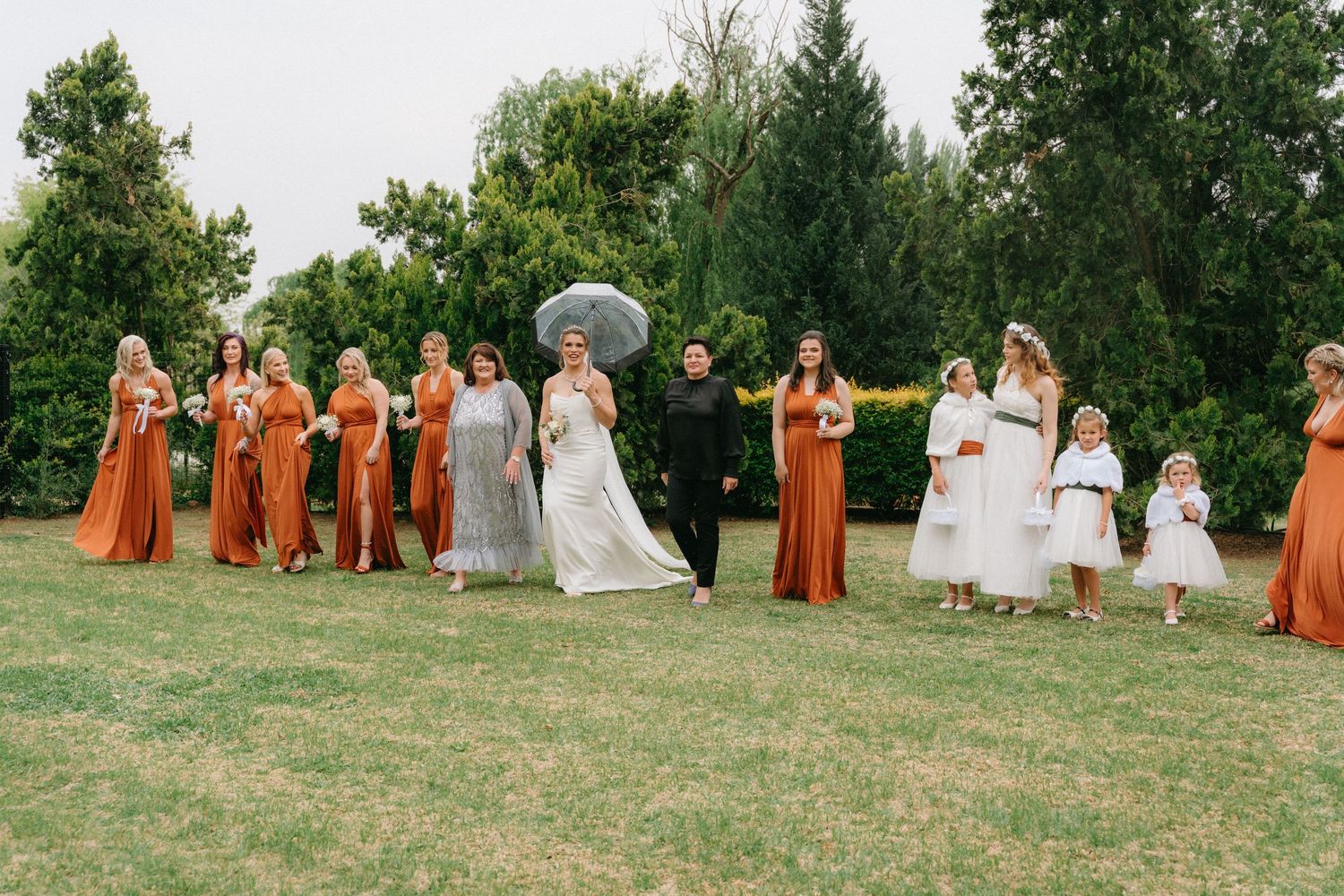 Wedding party poses on grassy lawn with bridesmaids in rust orange dresses.