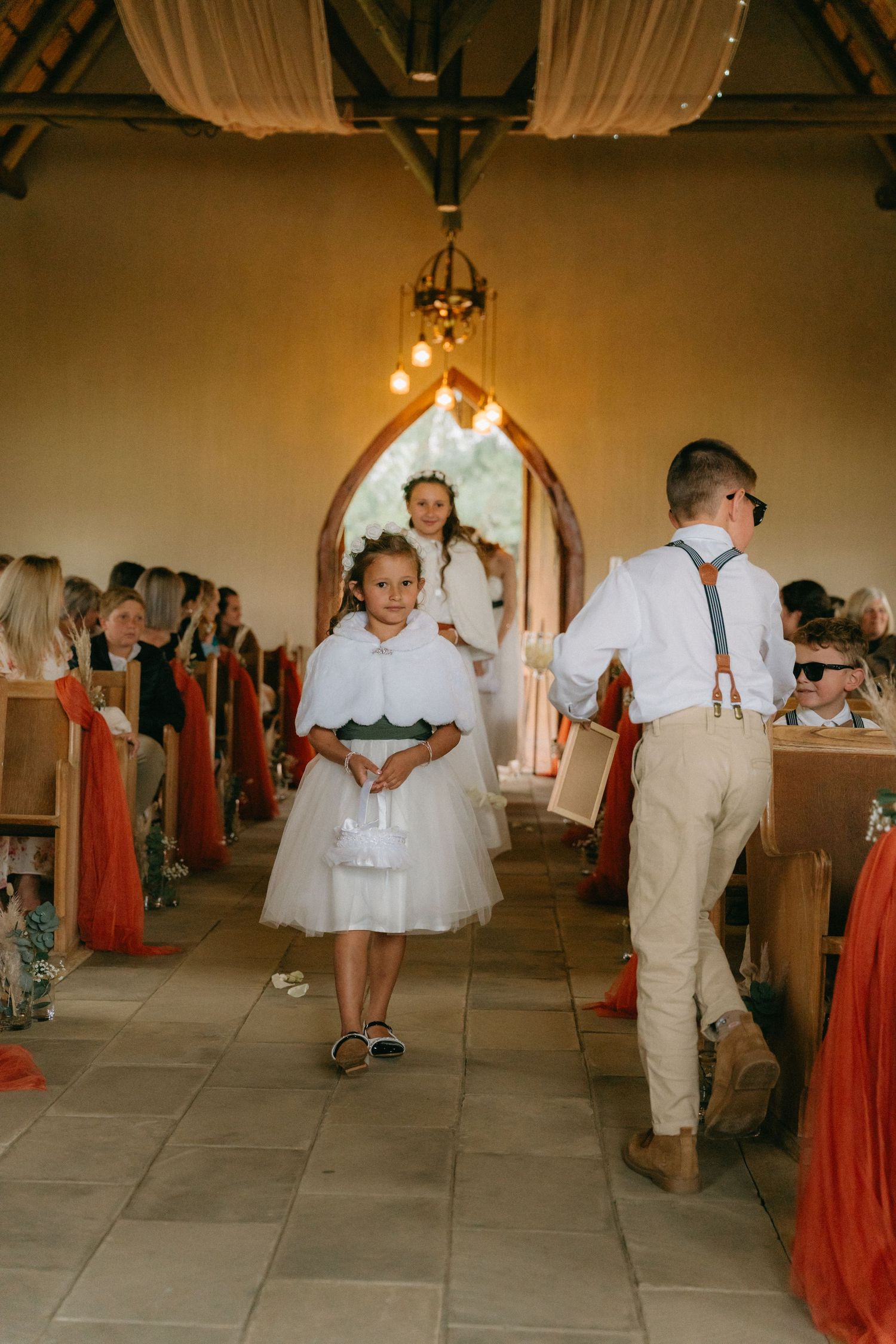Ring bearer and flower girl walking down the aisle in white outfits with red decor.