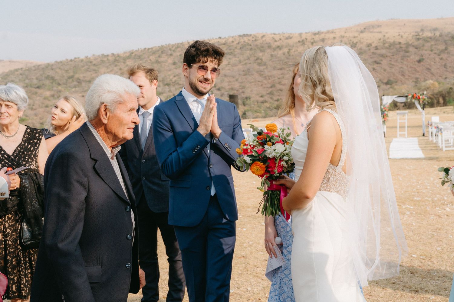 Wedding guests gather outdoors in a desert setting with mountains in the background during a celebration.