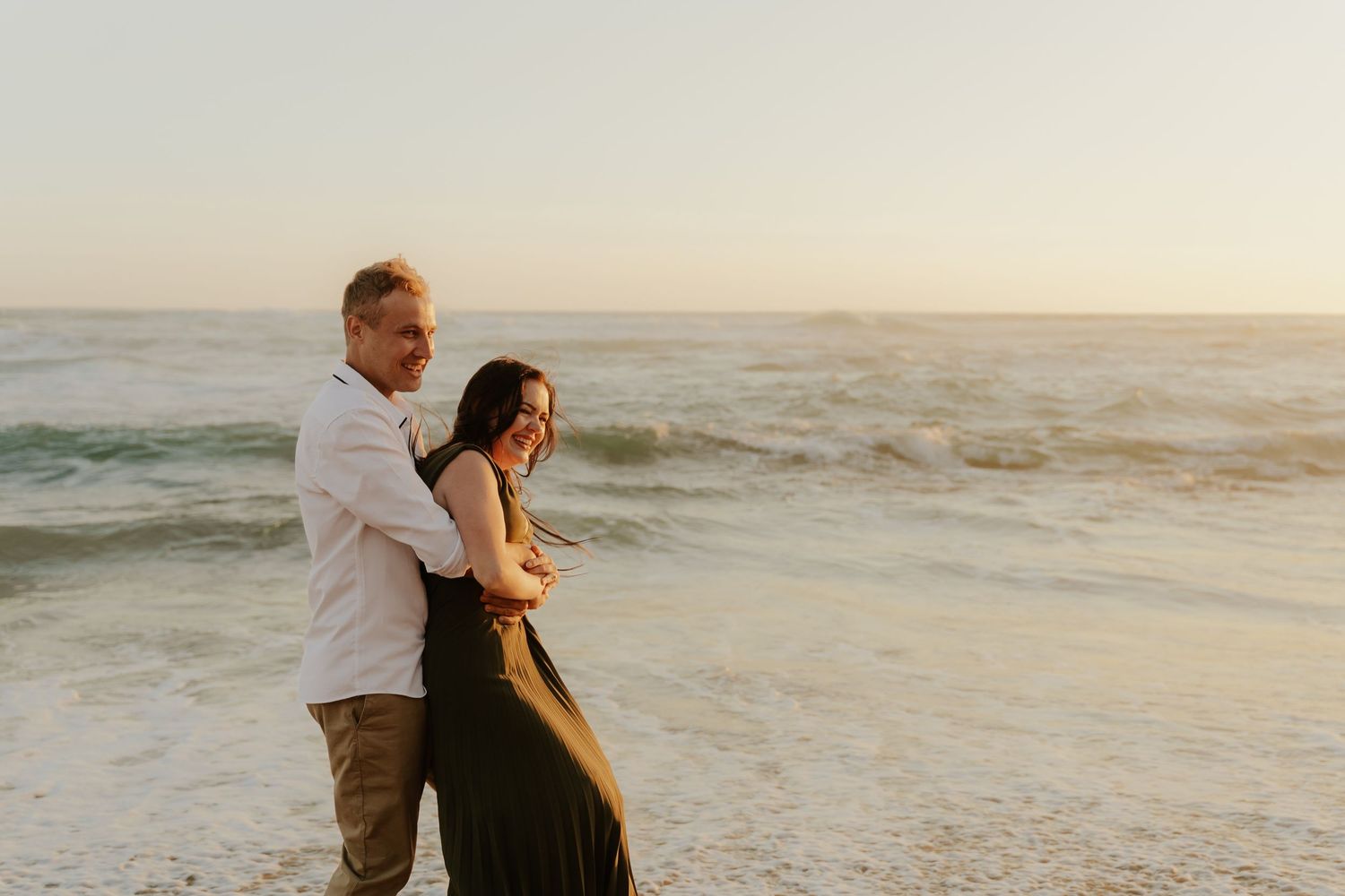 Couple embracing on beach at sunset while waves crash on shore in romantic seaside moment.