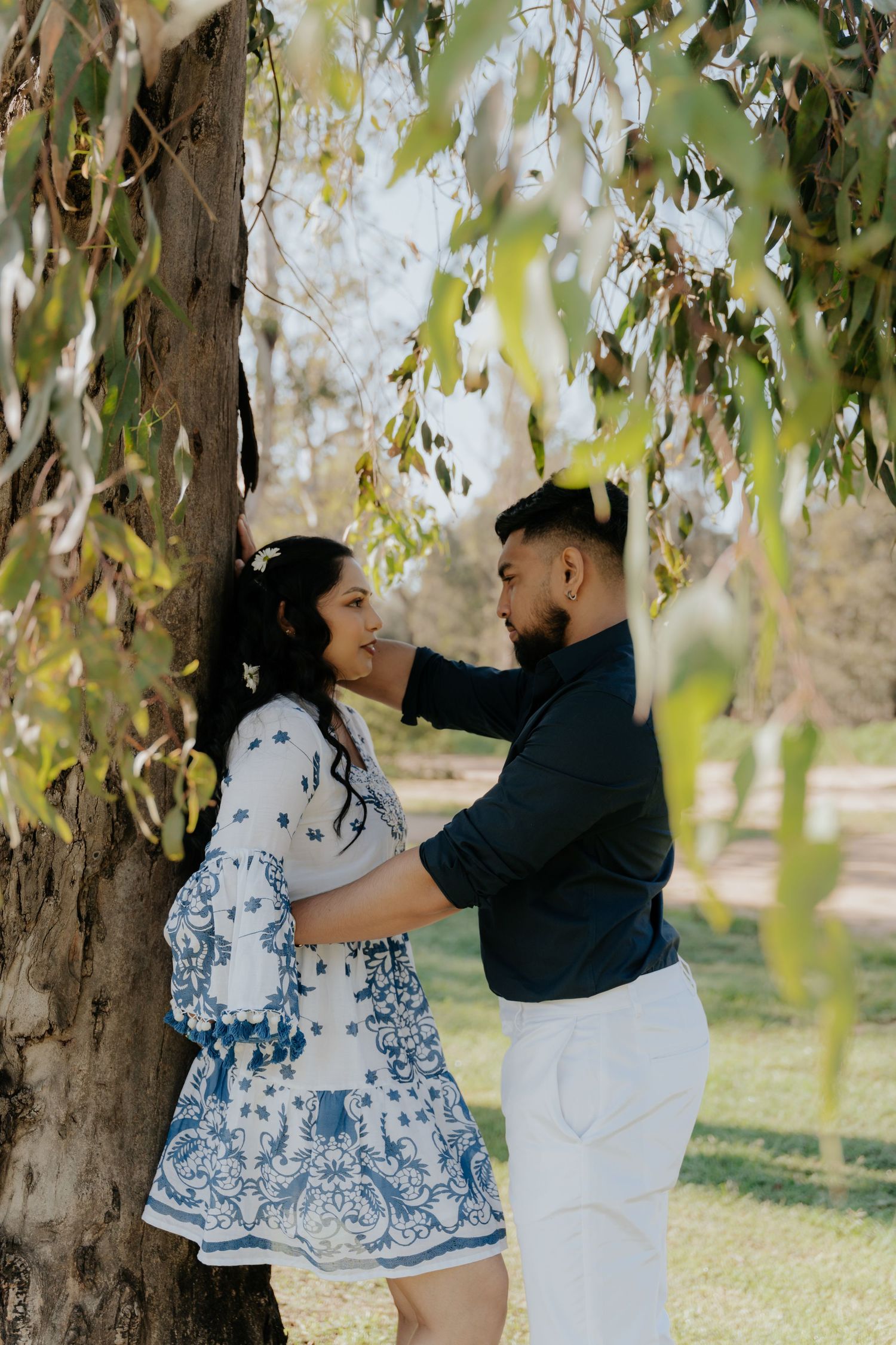 A couple shares romantic moments under a willow tree in a park setting, wearing blue and white outfits.
