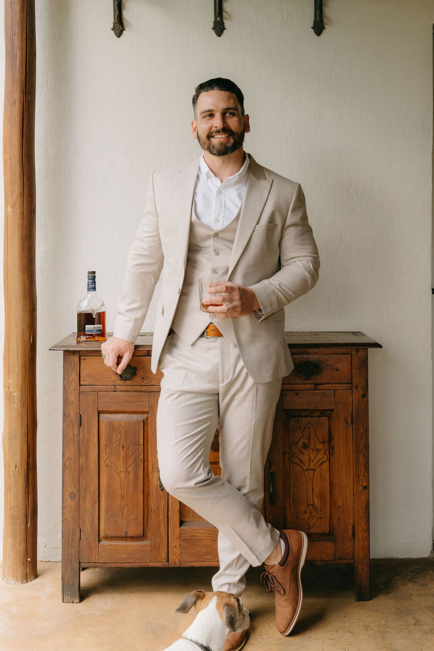 Groomsmen in beige suits relax with drinks against wooden furniture.