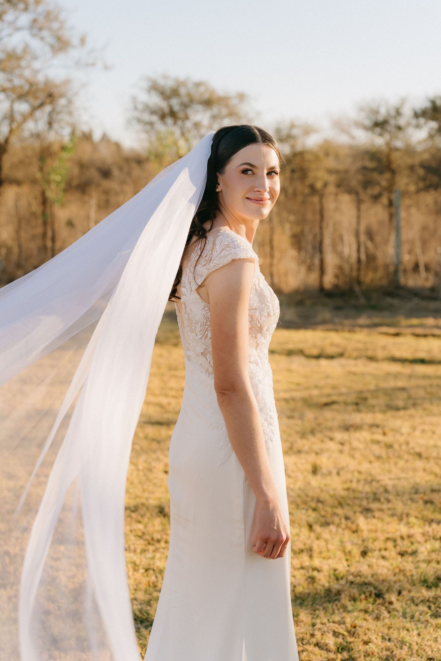 Flowing white wedding gown with lace details and long veil captured in golden sunlight.