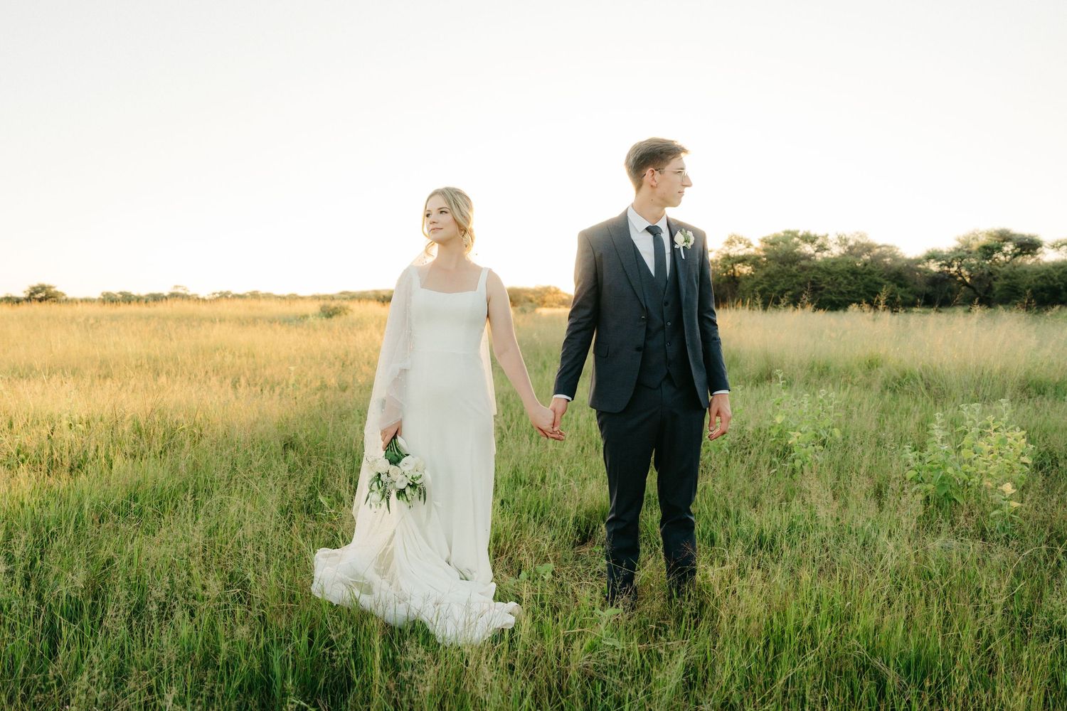 A couple stands together in a serene meadow at sunset wearing formal wedding attire.