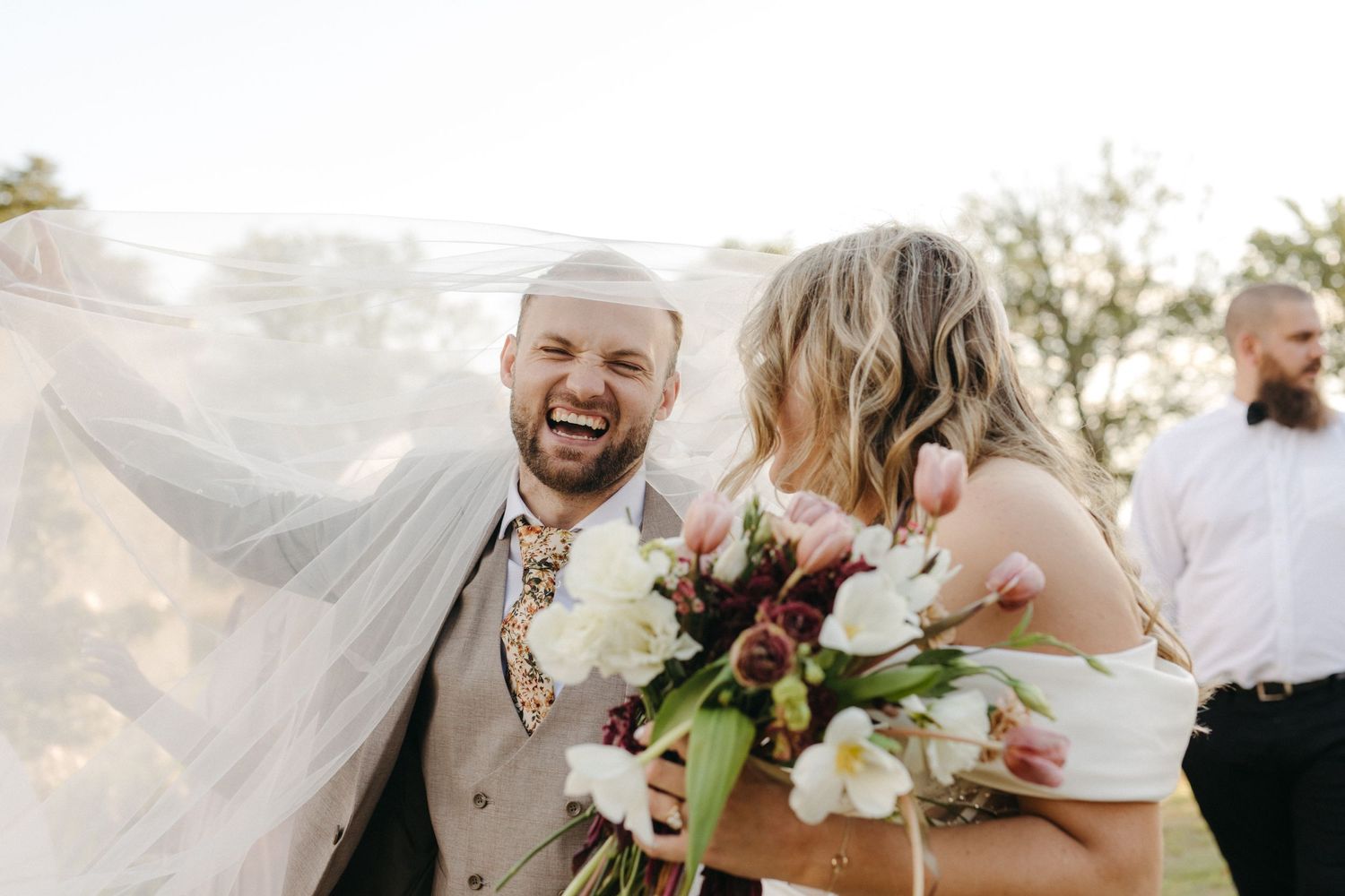 Joyful wedding couple laughing together with white veil and bouquet of flowers in sunlit outdoor ceremony.