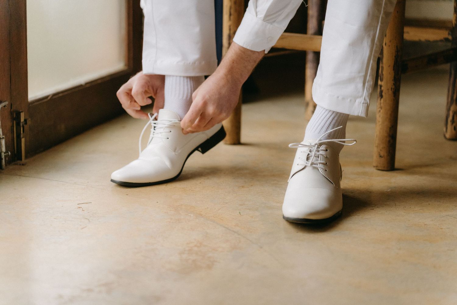 Close-up of groom tying white leather dress shoes on wooden floor.