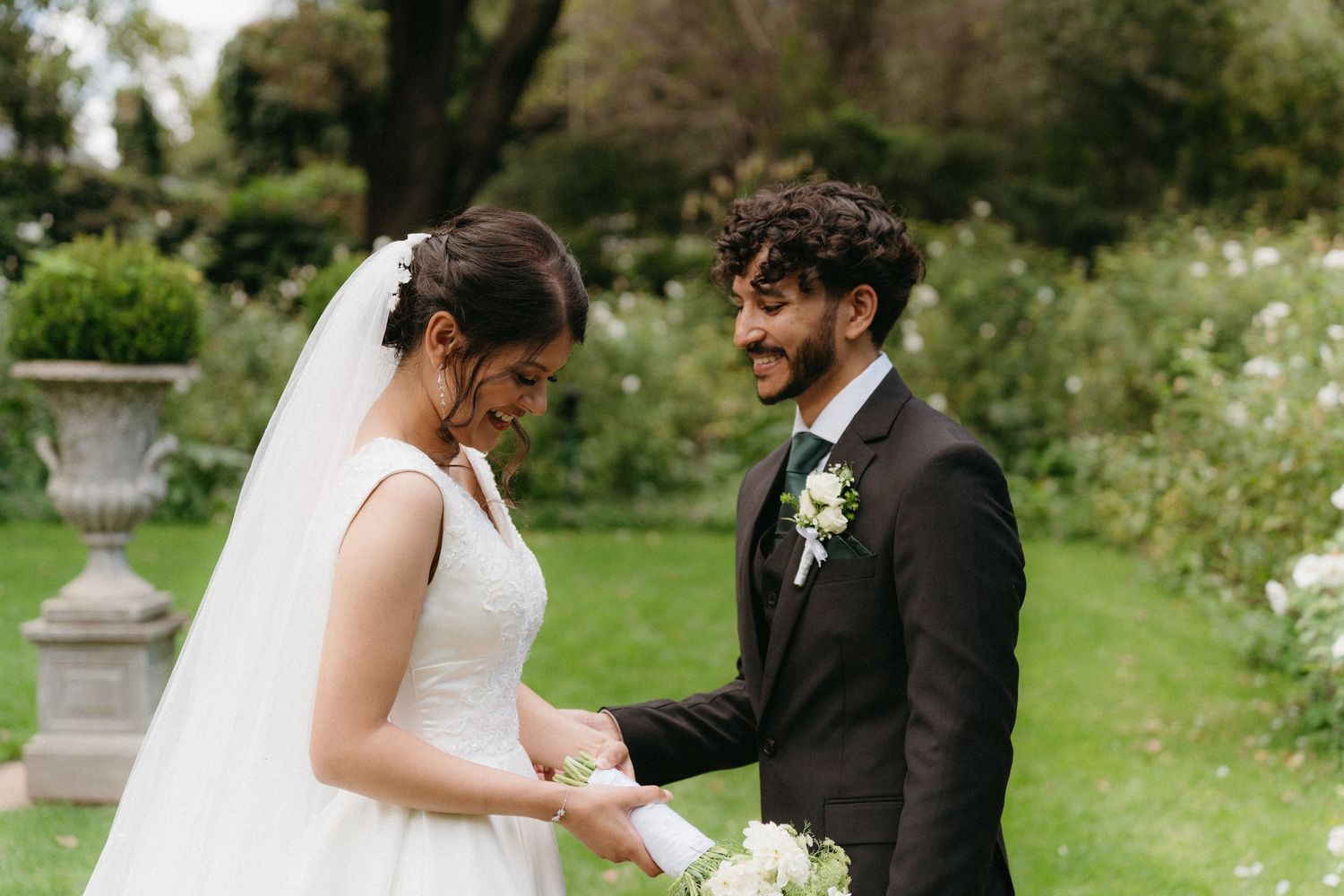 A joyful candid moment between a bride and groom laughing together in a garden setting with decorative urns.
