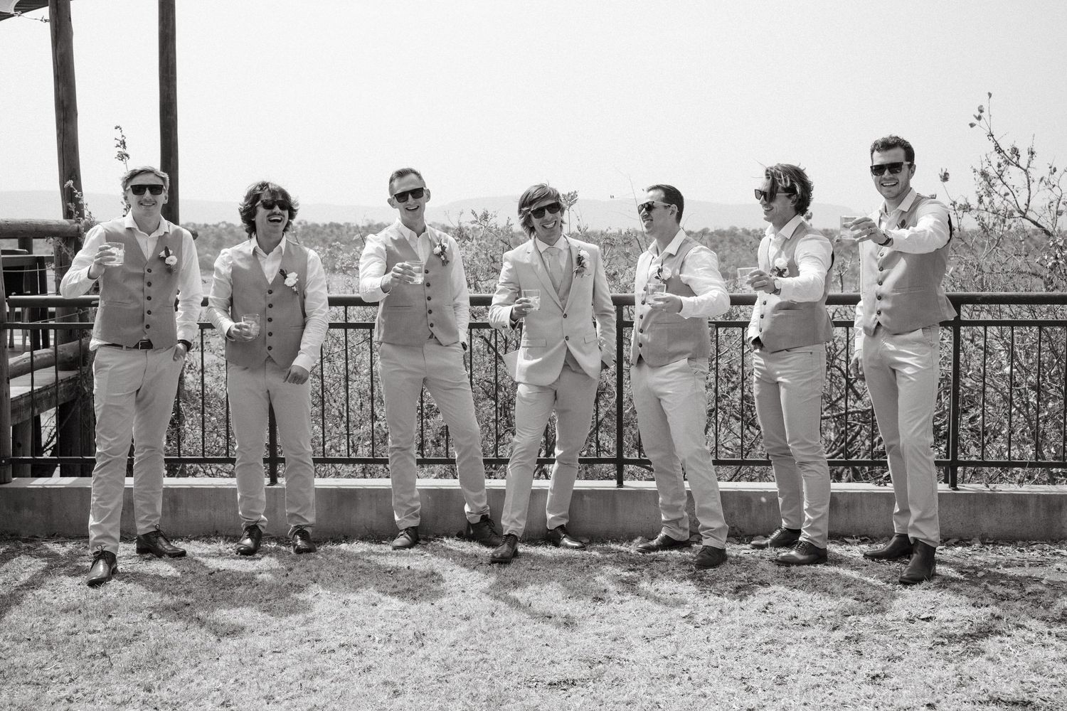 Groomsmen in light colored suits sit casually on a stone wall for a group photo.