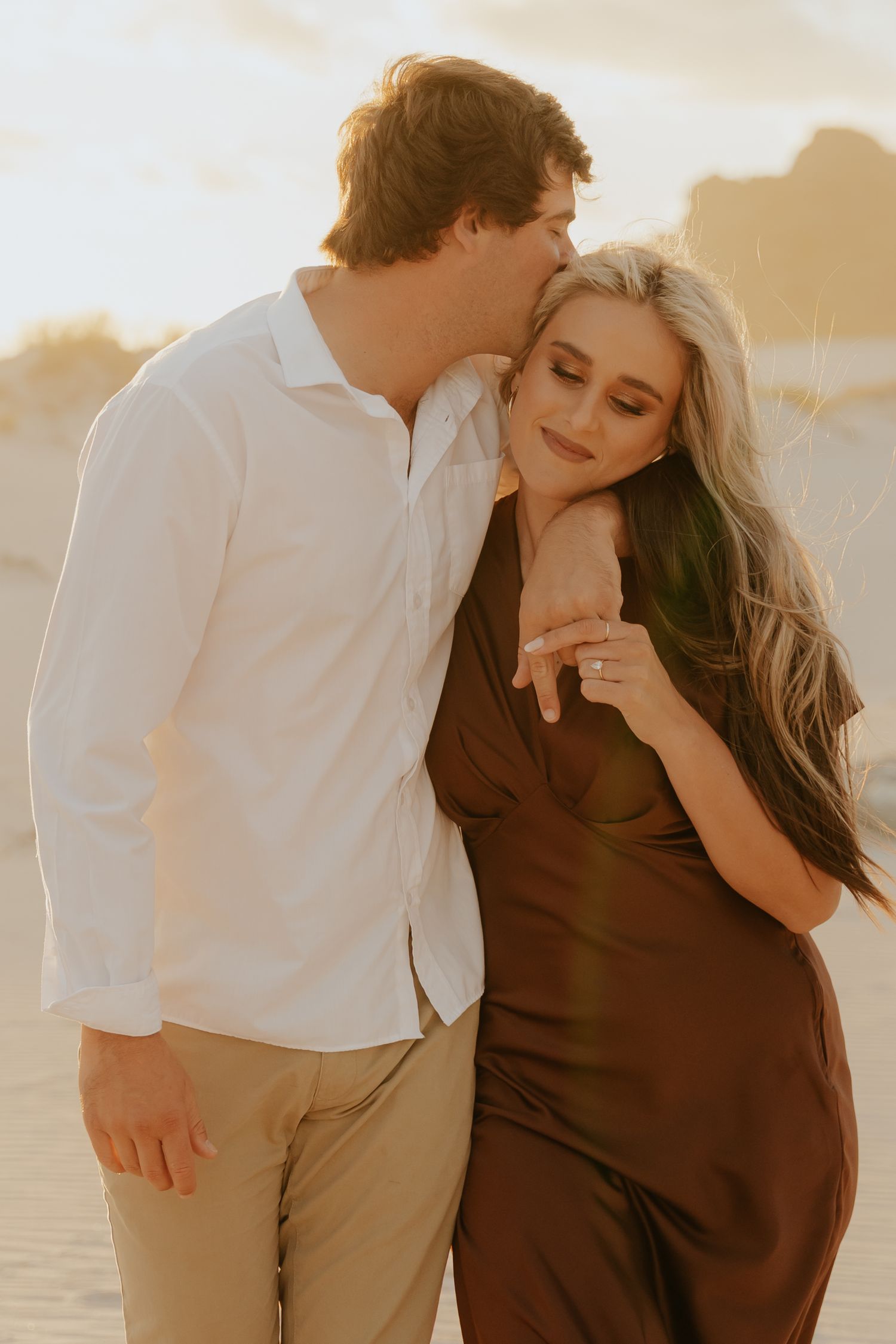 Couple sharing a romantic moment on sandy dunes at golden hour, woman in brown dress showing engagement ring.