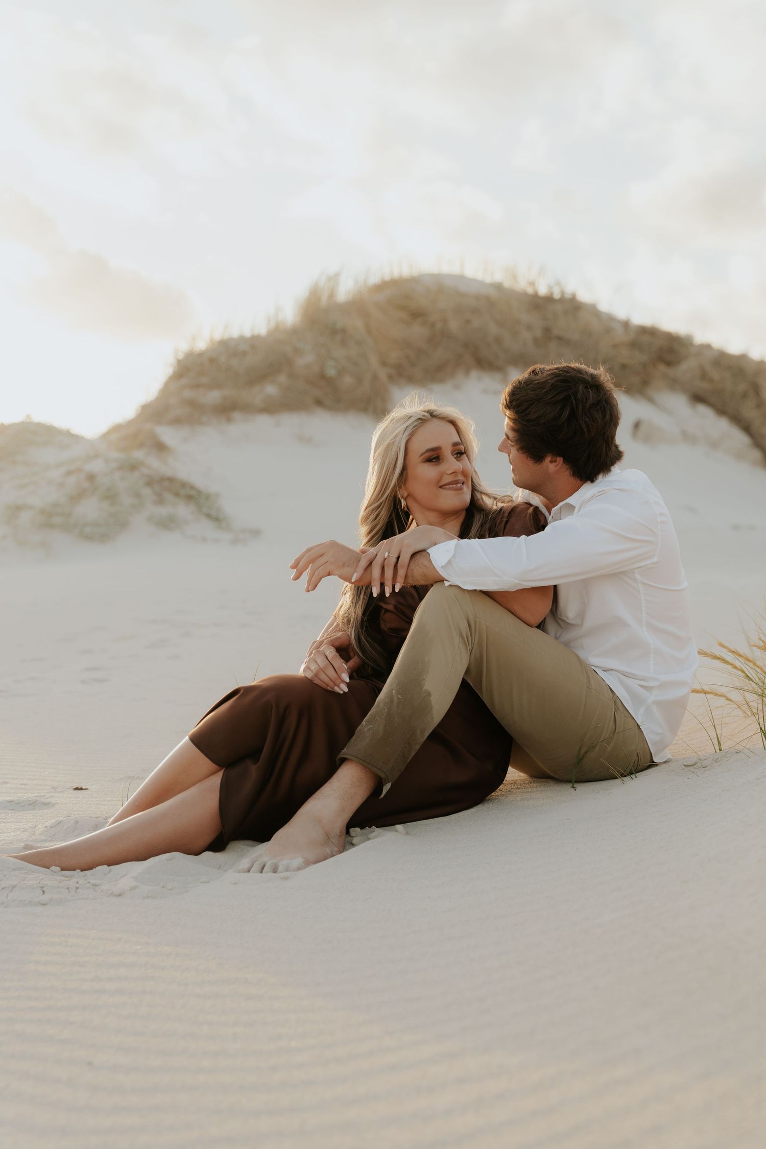 A couple sits barefoot on sandy dunes, the woman in a brown dress embracing a man in white, sharing a romantic moment.