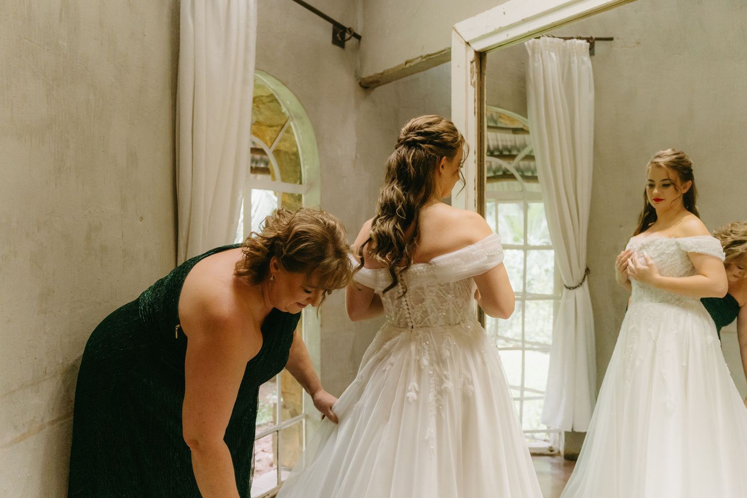 People in formal attire help with wedding dress preparations in an elegant dressing room.