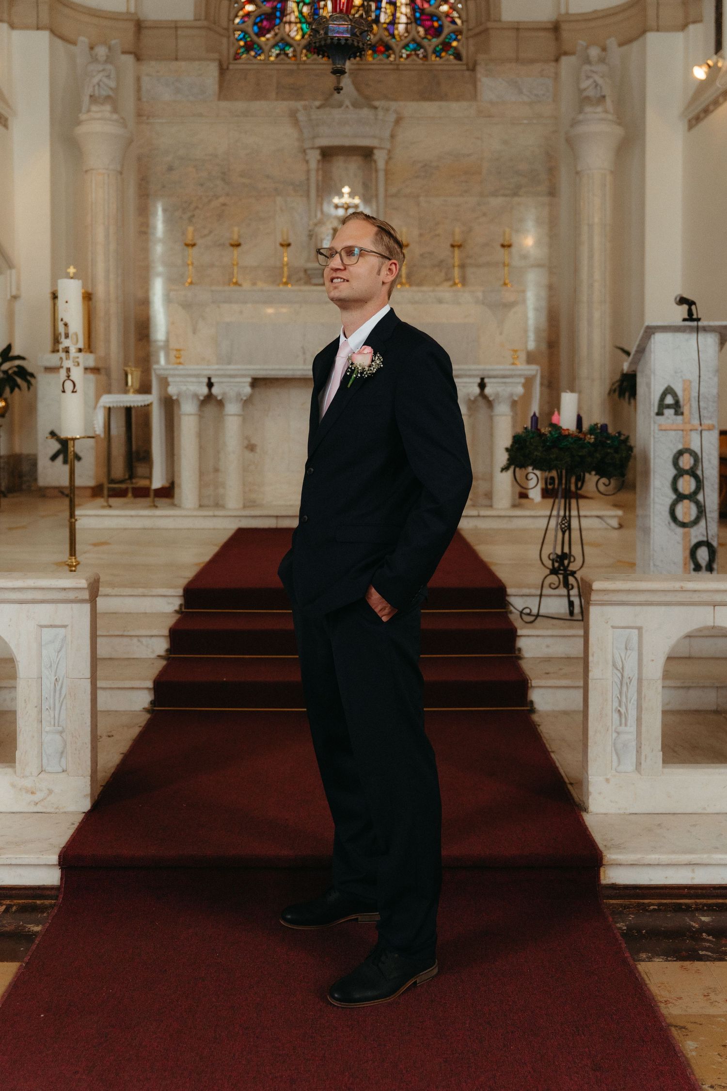 A groom in a navy suit and glasses poses for a portrait at Nazareth House Chapel in Pretoria