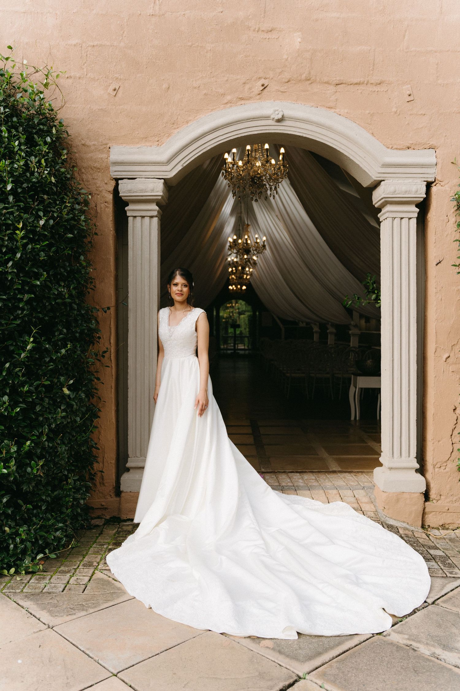 Bride in stunning white gown with long train stands in arched doorway with chandelier at Oakfield Farm in Johannesburg