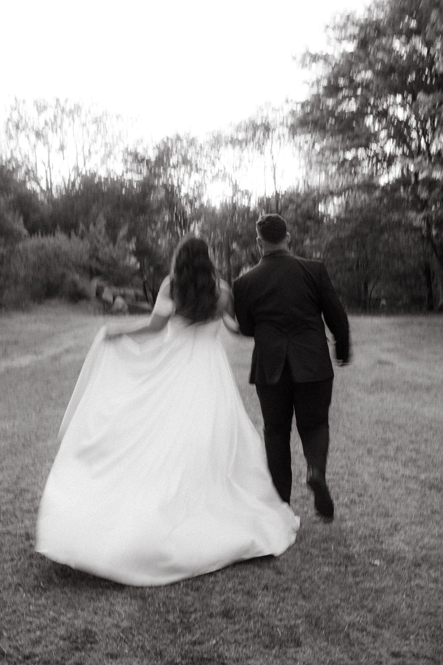 Black and white wedding photo of a couple walking away together on a grassy path surrounded by trees at dusk.