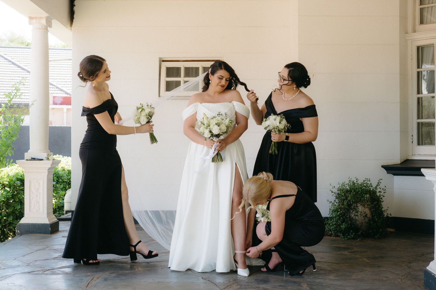 A bridal party shares a candid moment on a white columned porch, with bridesmaids in black and the bride in white.