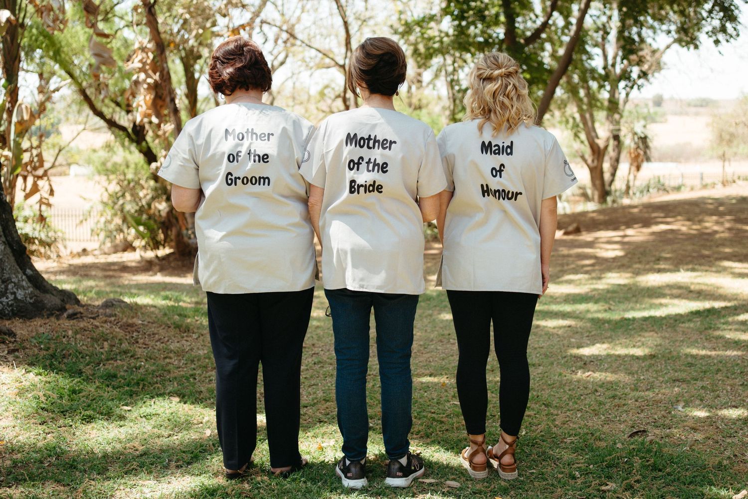 Three matching white t-shirts with wedding party roles printed on the back viewed from behind.