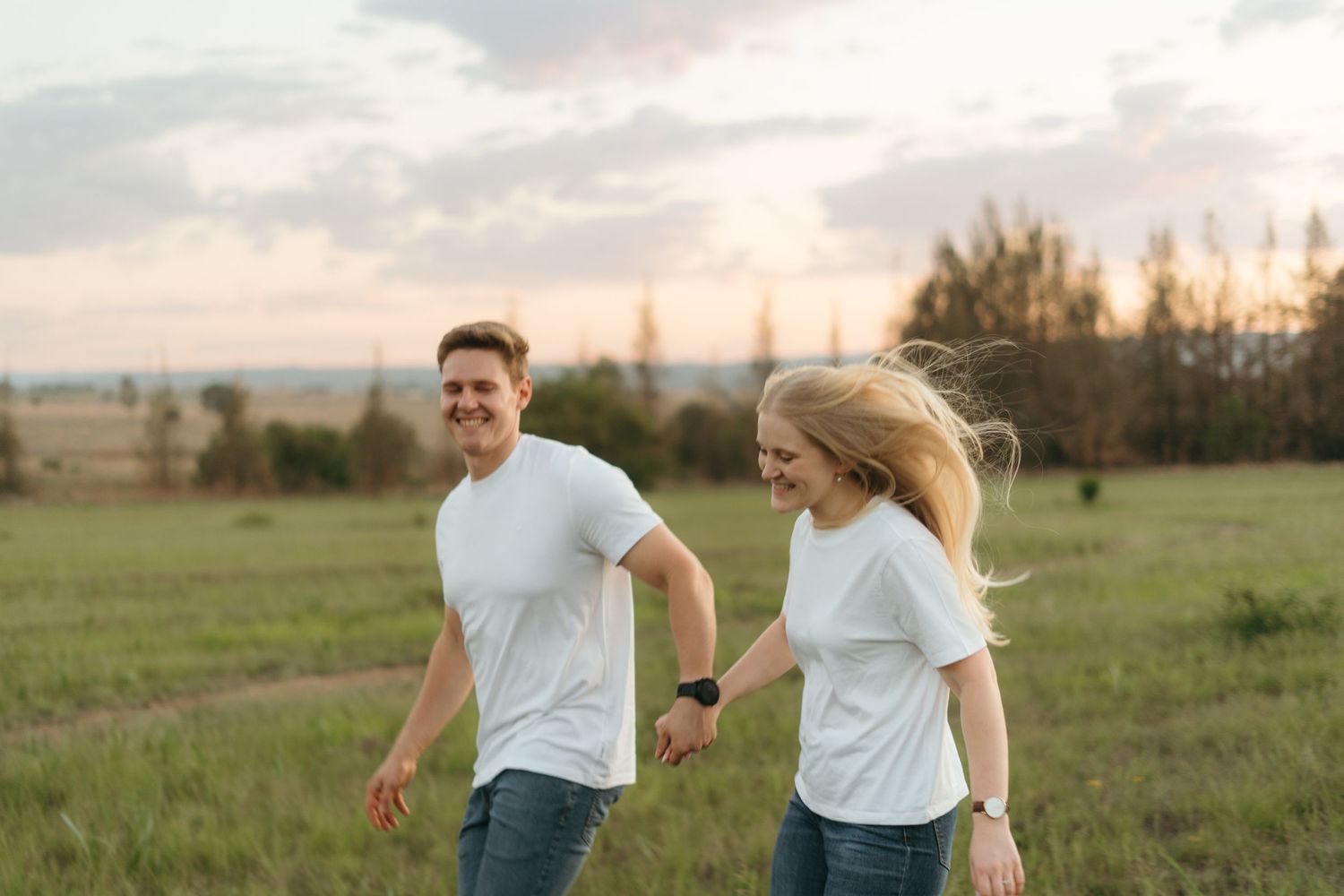 Joyful couple in matching white outfits walking through a green field at dusk.
