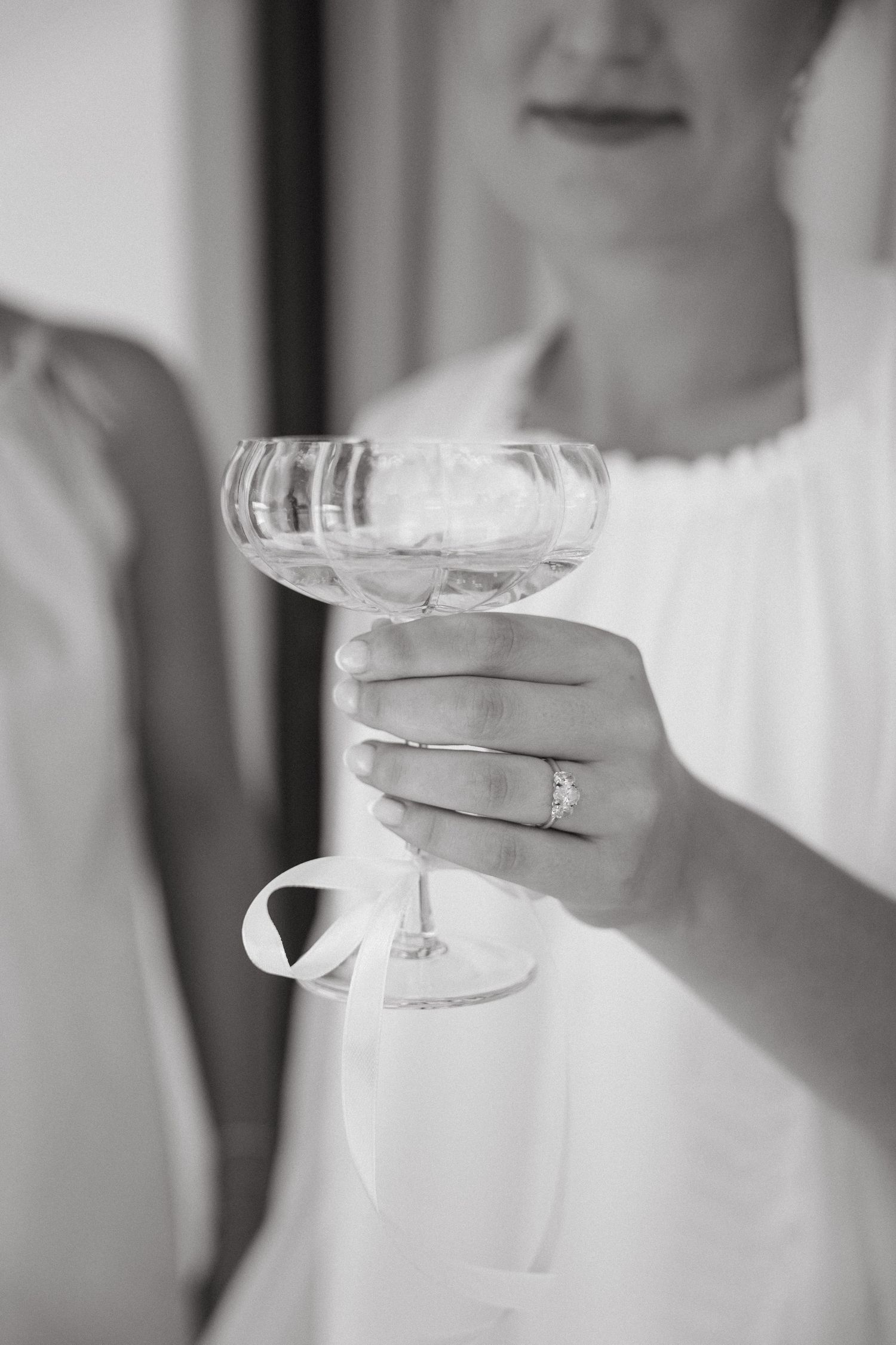 A hand wearing an engagement ring holds a champagne coupe glass in an elegant black and white photograph.