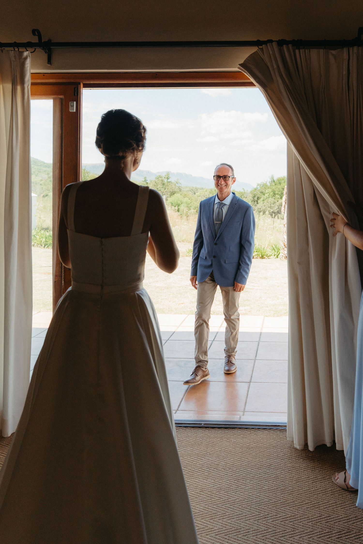 A wedding first look moment as a couple sees each other through an open doorway with sunlit outdoor scenery behind.